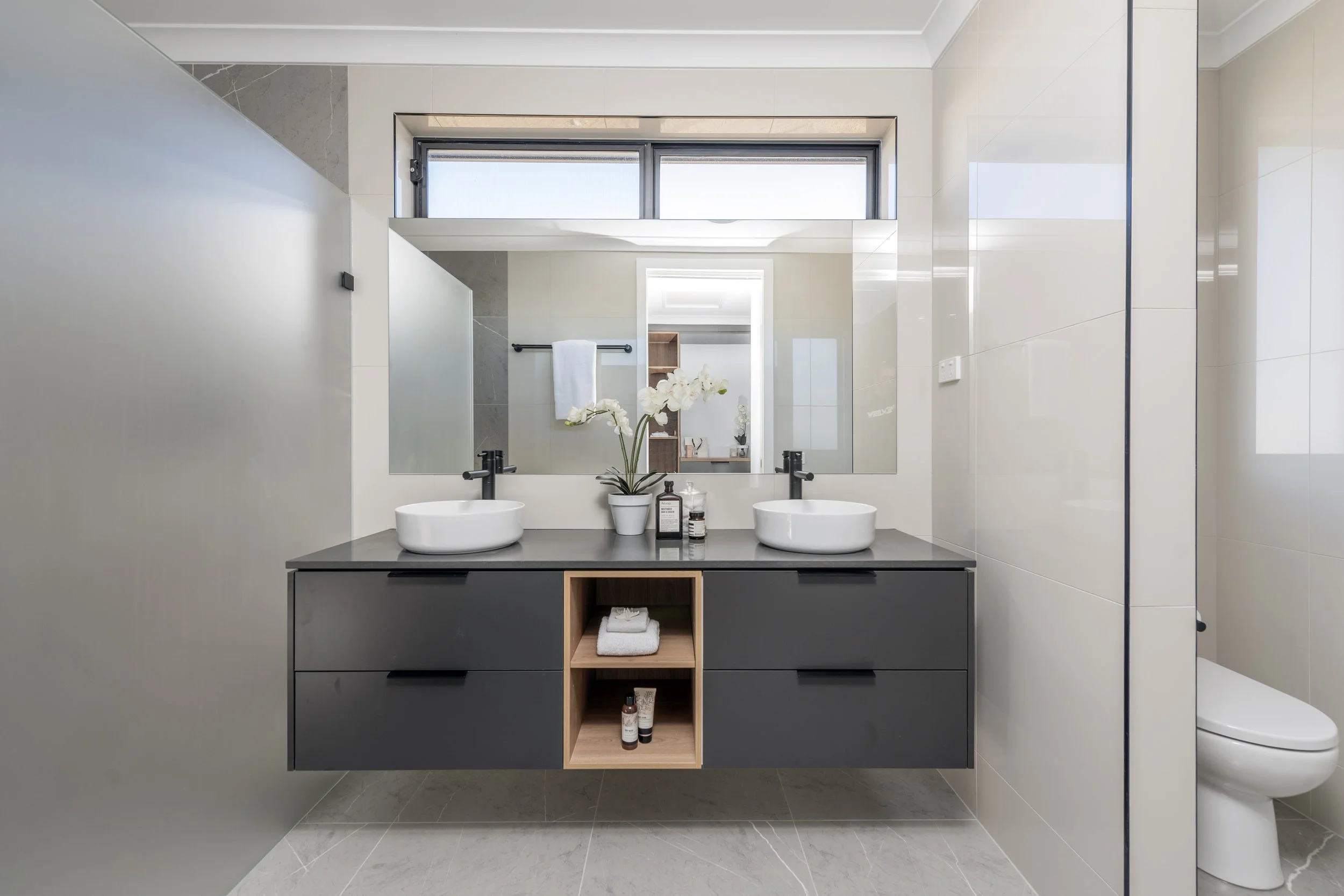 Modern bathroom with double vessel sinks, large rectangular mirror, gray vanity with open shelving, light-colored tiled walls and floors, and a window at the ceiling.
