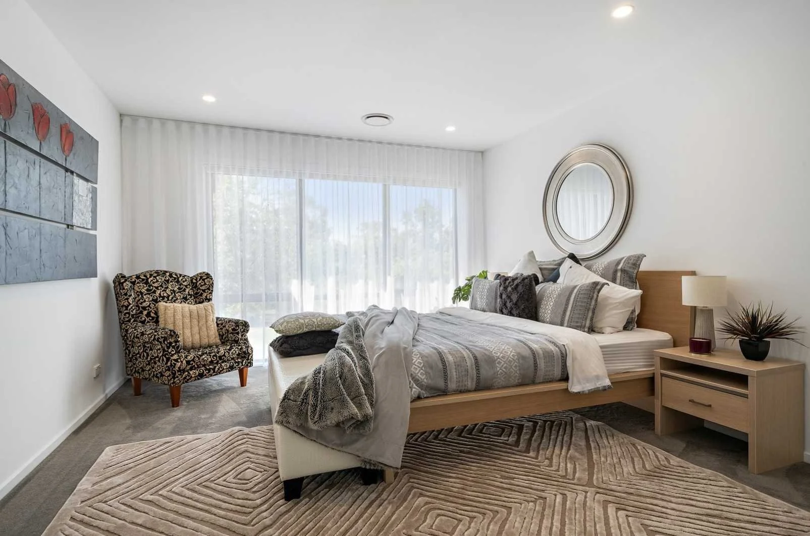 A bedroom with a wooden bed, nightstand, and mirror, featuring patterned pillows, a lamp, and artwork, with a large window and sheer curtains.