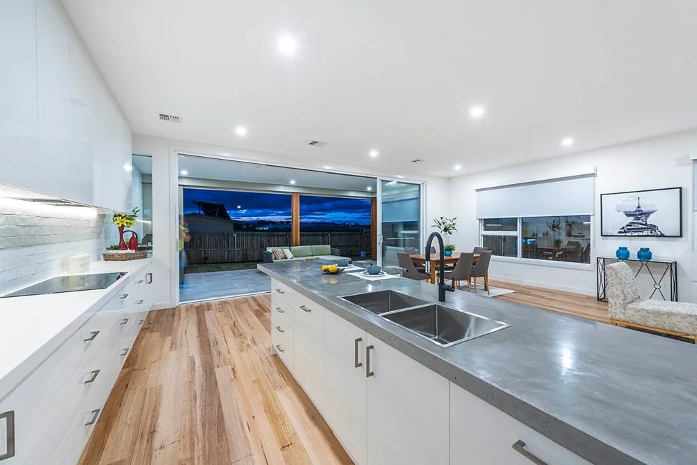 Modern open-concept kitchen and dining area with white cabinets, wooden floors, and a large sliding glass door leading to a patio with outdoor seating, nighttime sky visible outside.