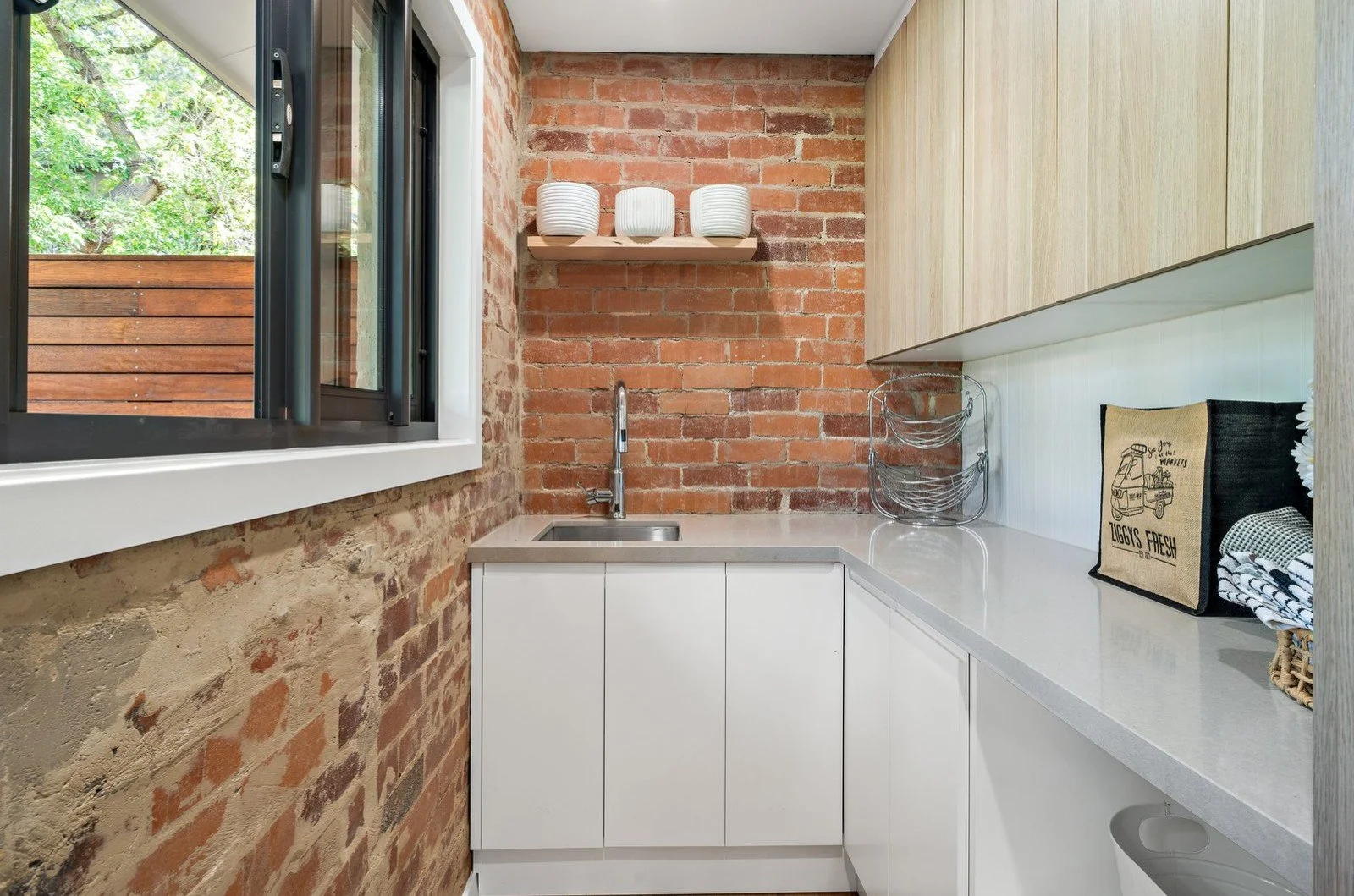 Small kitchen area with exposed red brick walls, a window showing greenery outside, white lower cabinets, a white countertop, a minimalist sink with a modern faucet, and shelves with white bowls. A black cloth bag on the counter reads "LIGGYS FRESH,"