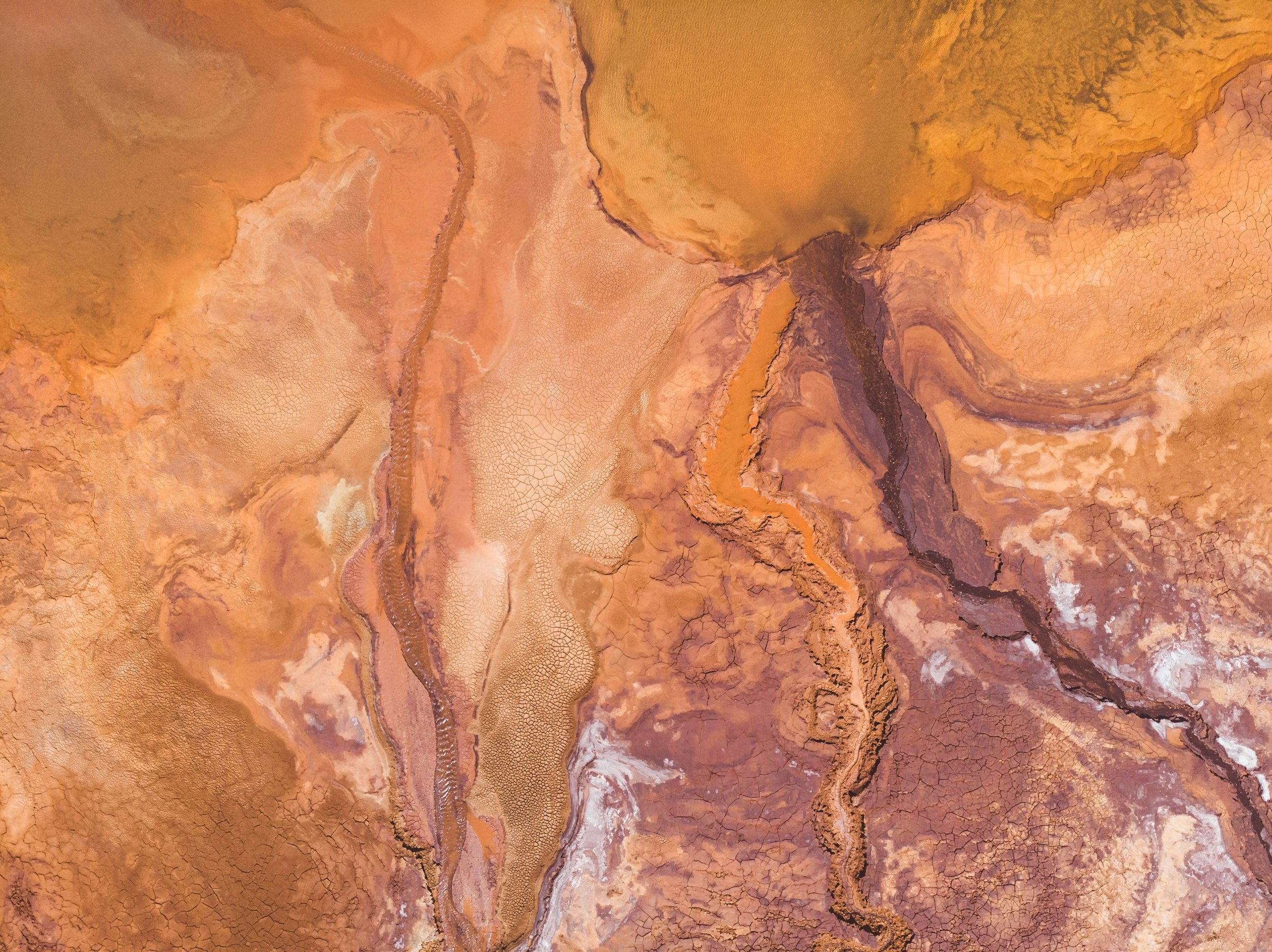 Aerial view of colorful geothermal features with mineral deposits and hot springs in shades of orange, brown, white, purple, and yellow.