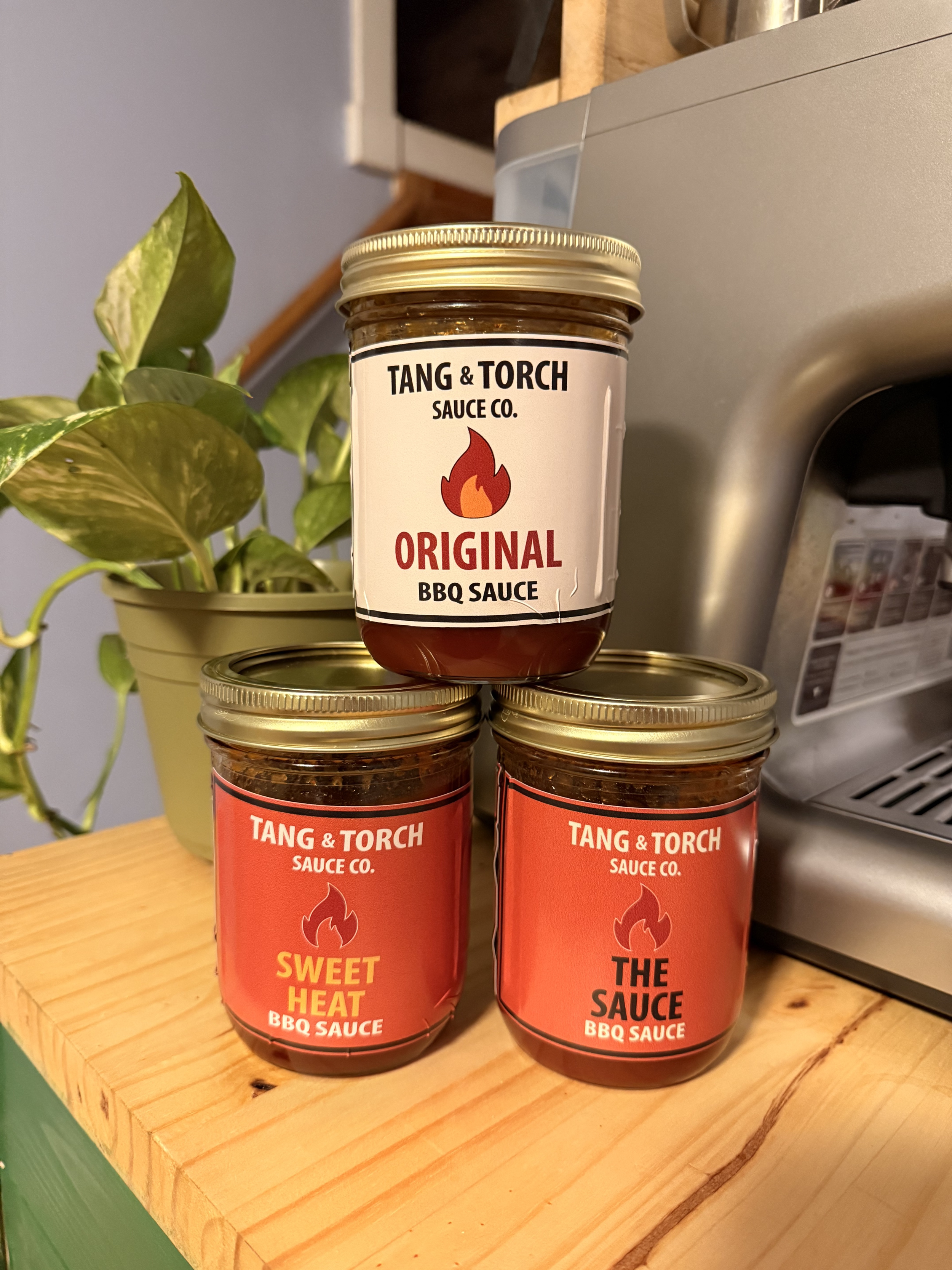 Three jars of BBQ sauce from Tang & Torch Sauce Co. placed on a wooden surface, with a plant in a yellow pot and a stainless steel appliance in the background.