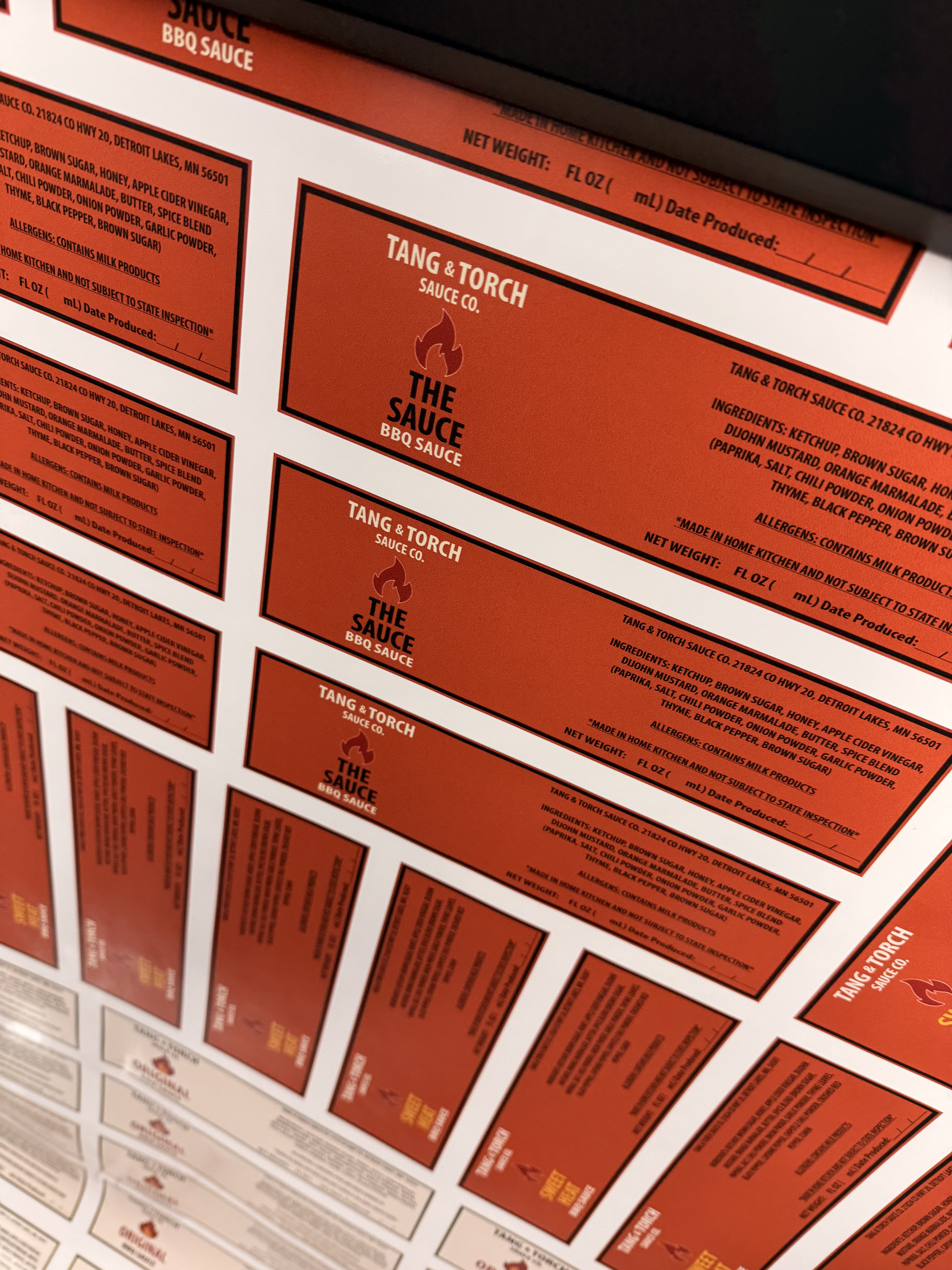 Close-up of multiple red and white food labels, prominently featuring 'Tang & Torch Sauce Co.' and 'The Sauce BBQ Sauce', with ingredients and allergy information.