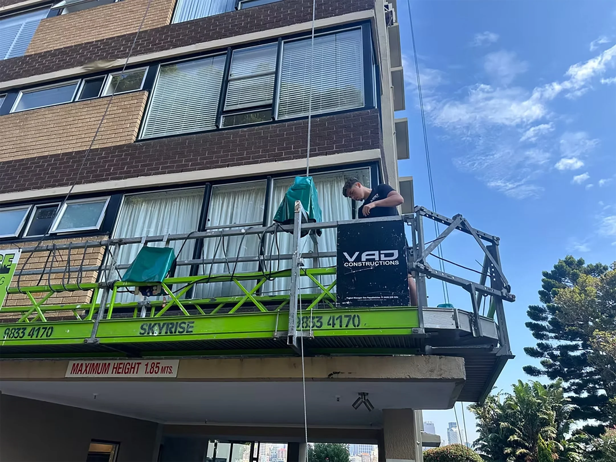 A man working on a balcony of a multi-story building with blue sky and clouds in the background. The balcony has a green and silver scaffolding with a sign that says "SKYRISE" and a maximum height warning. The building's exterior has brick and window features.