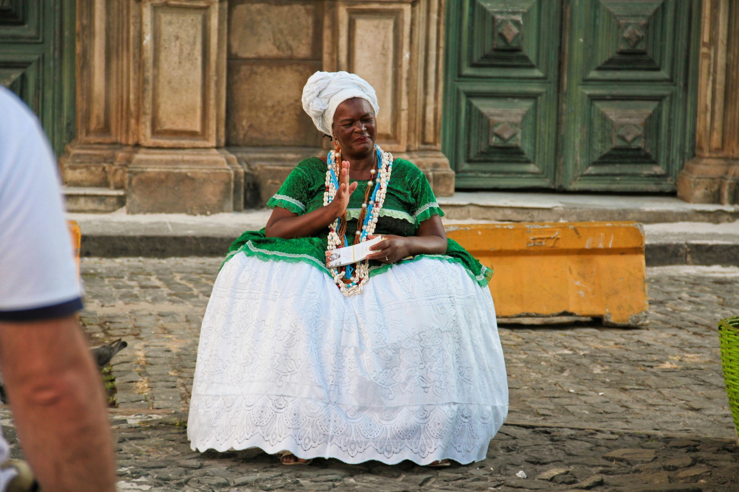 A woman dressed in traditional Bahian attire, wearing a green blouse and a white embroidered skirt, sitting on the cobblestone street.