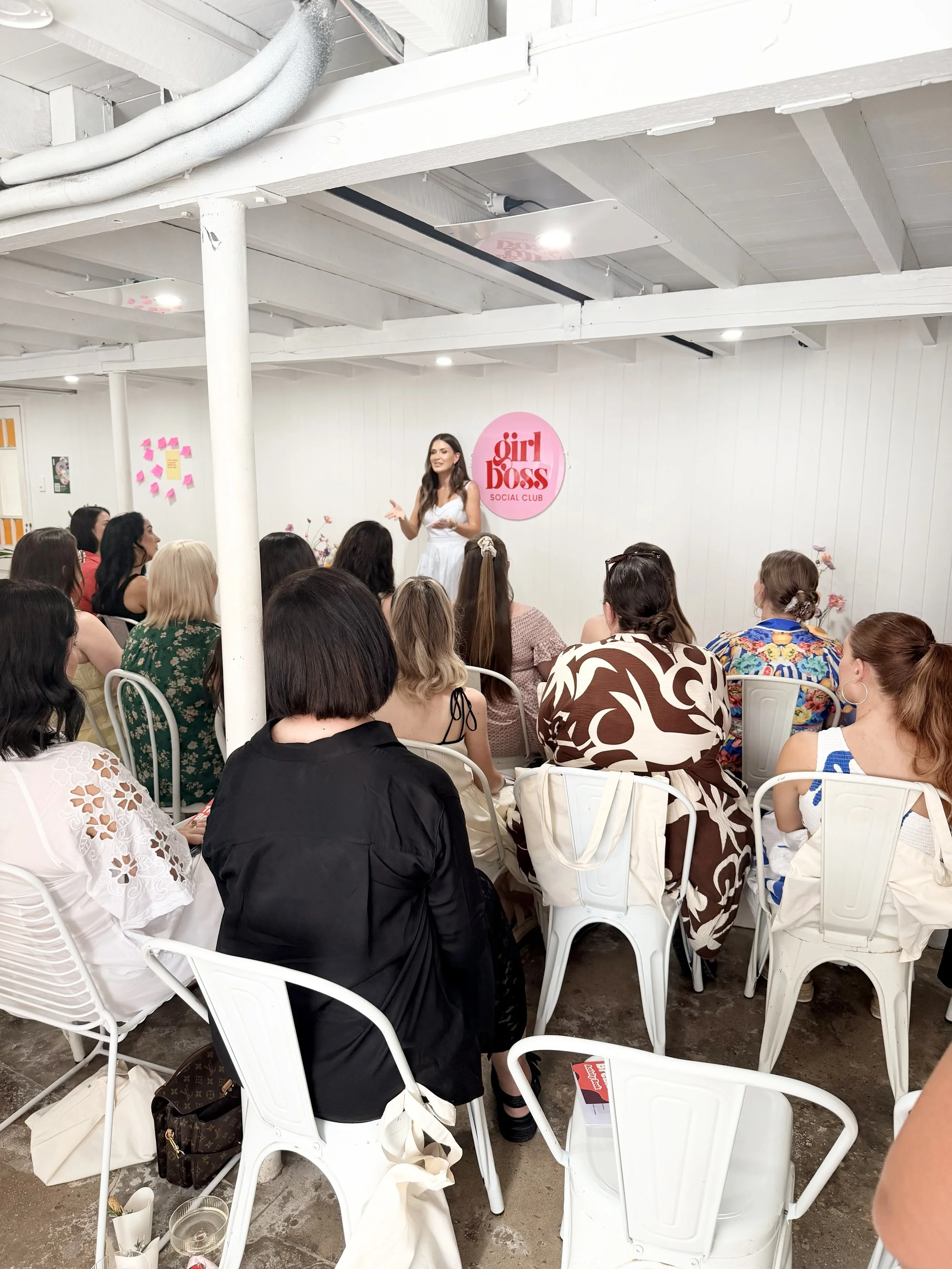 A woman giving a presentation or speech to a group of women in a white indoor space at a social club event called girl boss. The women are seated on white chairs, some with bags or belongings beside them, and the presenter is standing in front of a pink sign that reads 'girl boss'.