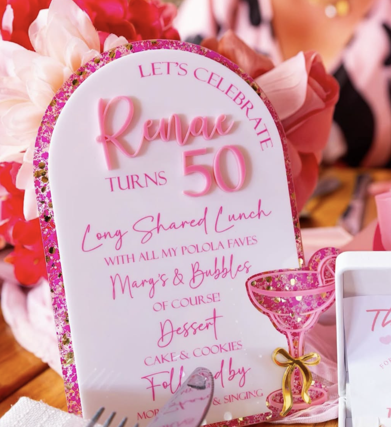 A sign celebrating Renae's 50th birthday with pink and white decorations, pink flowers, and a pink champagne glass.