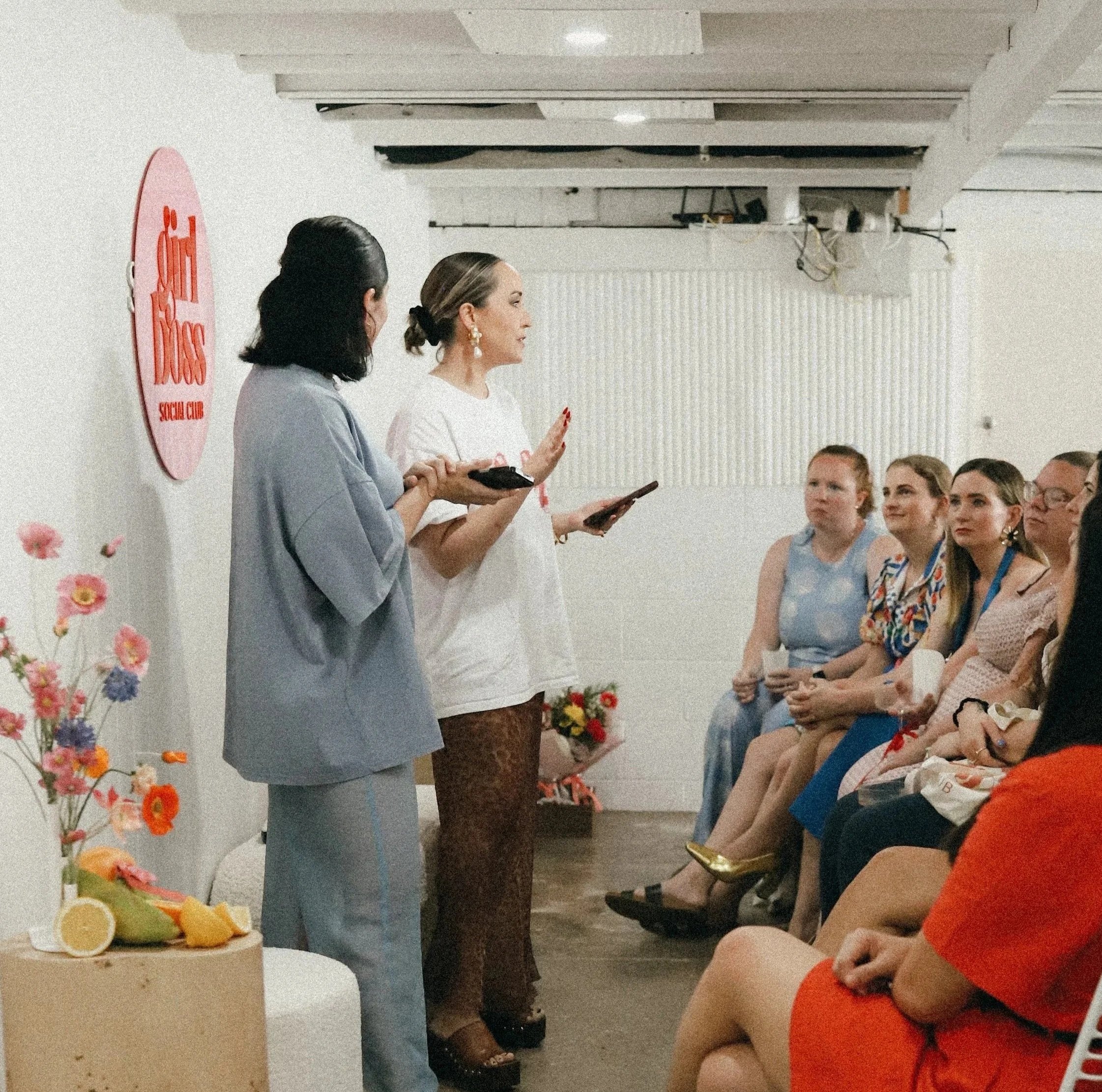 Two women standing and speaking in front of a seated audience at a social club event. One woman is dressed in a gray suit, and the other in a white t-shirt and leopard print pants. A group of women are sitting and listening attentively. Decorations include flowers and a hanging sign that says 'Flii Flii Social Club'.
