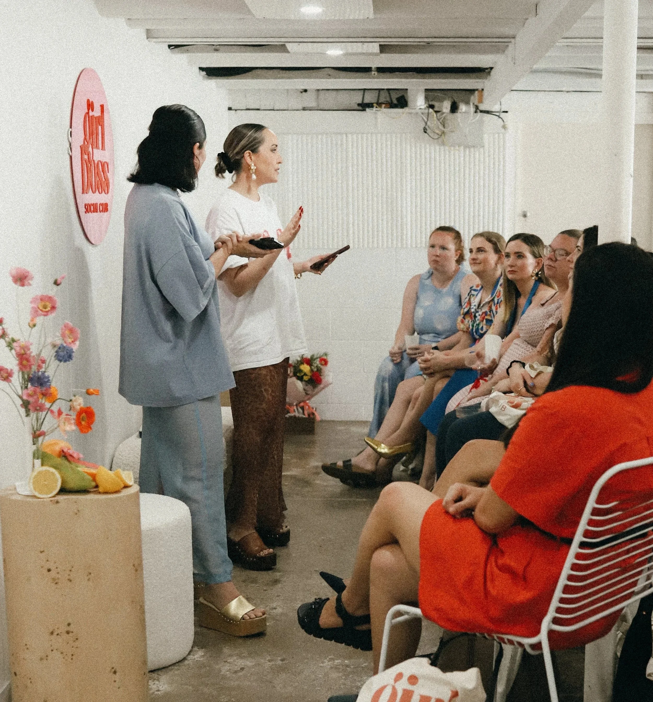 Two women giving a presentation to a seated audience in a room with white walls and exposed ceiling beams. One woman is holding a paper, and the other has a phone. The audience includes women in colorful dresses and casual clothing, some with notebooks or bags. Floral decorations and a sign reading 'Sex Dogs Social Club' are visible.