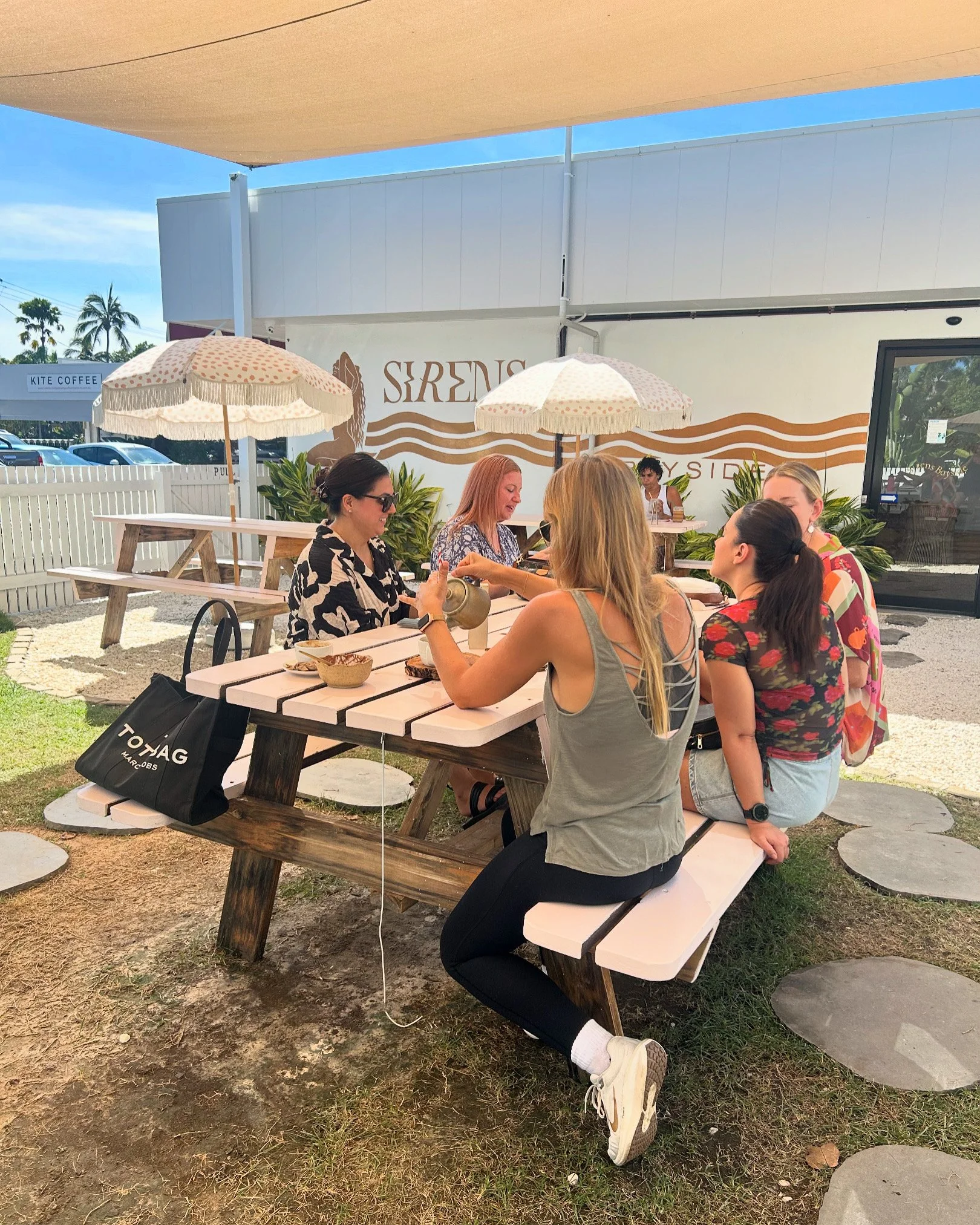 A group of five women sitting at a picnic table outside, enjoying a meal and conversation under umbrellas at a restaurant or cafe with a sign reading 'SIRENS' in the background.