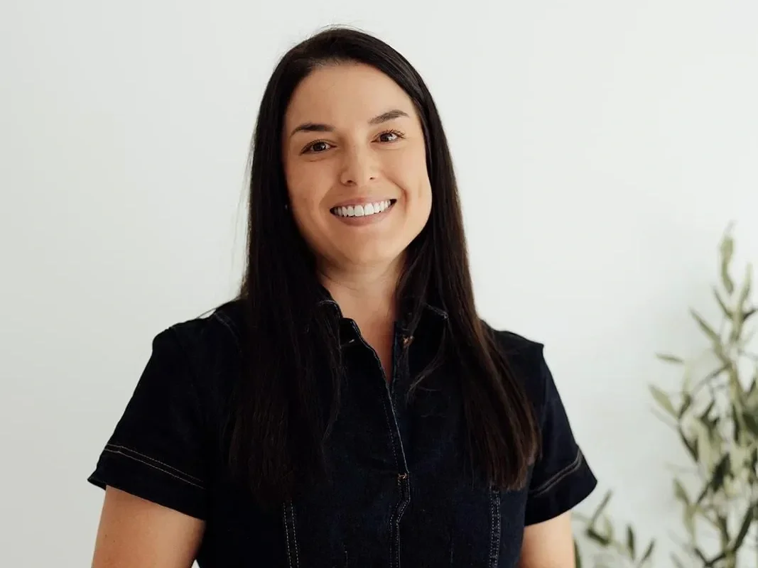 A woman with dark hair smiling, wearing a black shirt, standing indoors next to a plant.