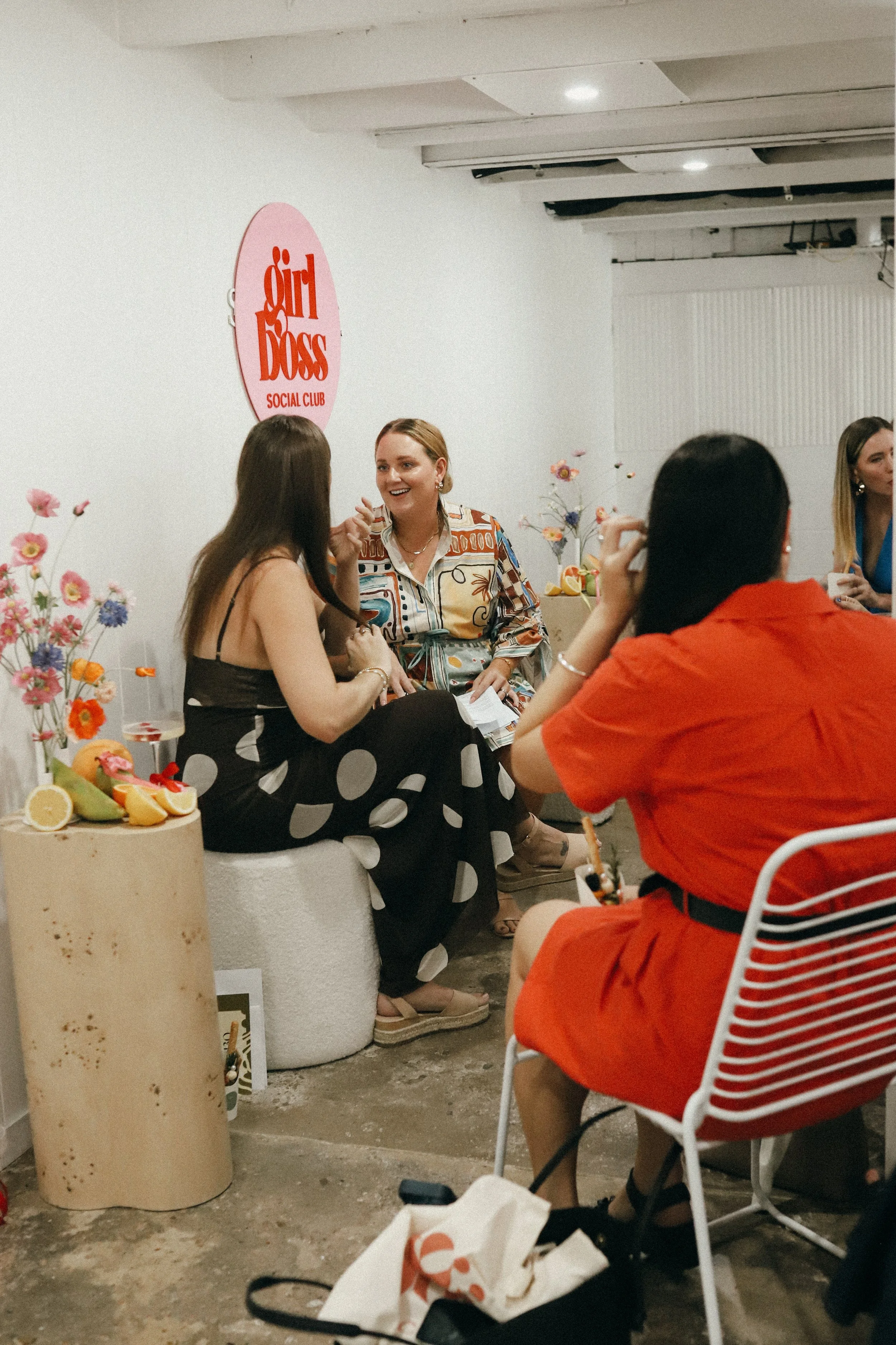 Group of women talking and socializing at a brunch or outdoor event, decorated with flowers and fruit. Pink sign on the wall reads 'Girl Boss Social Club'.