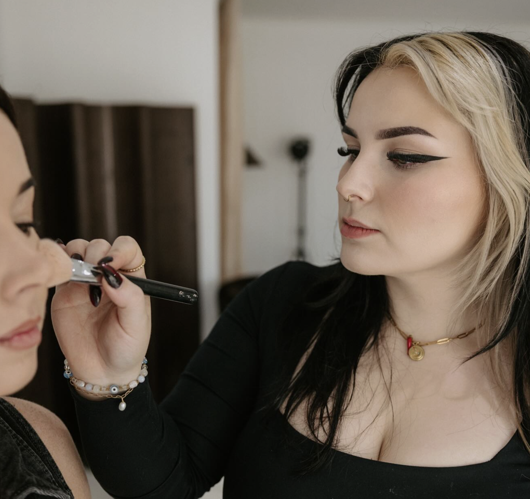 A makeup artist applying eye makeup to a woman with blonde and black hair, wearing a black top, gold necklace, and multiple bracelets.