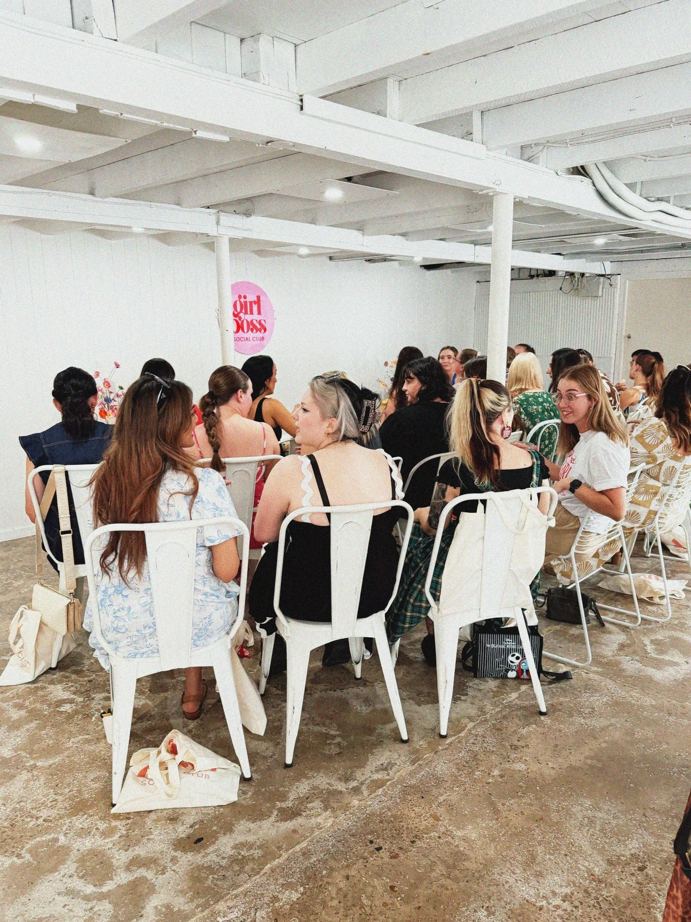 Group of women sitting around a table at a social club event with a 'Girl Boss Social Club' sign on the wall.