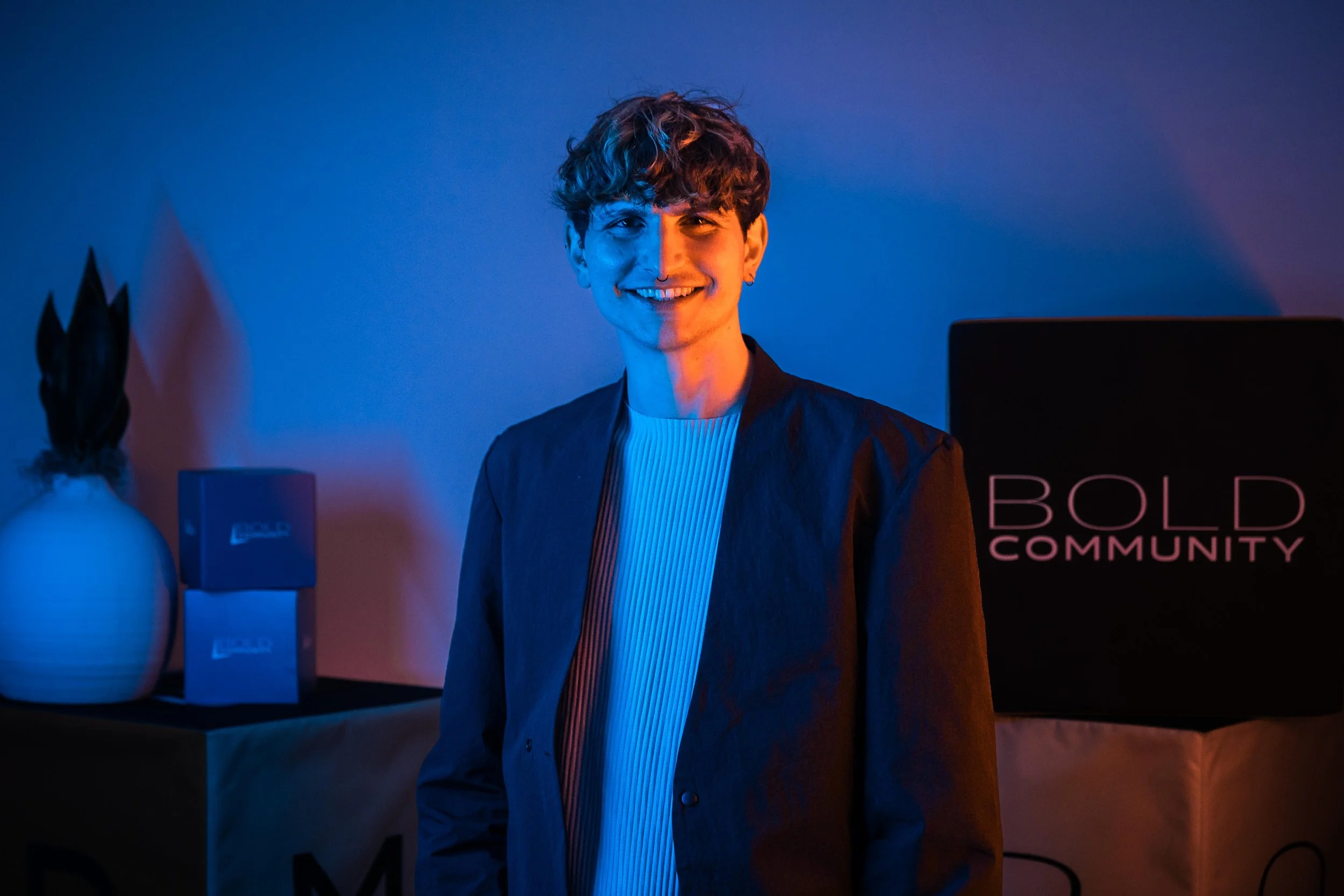 Young man with curly hair smiling, standing in front of a dark background with blue and orange lighting.