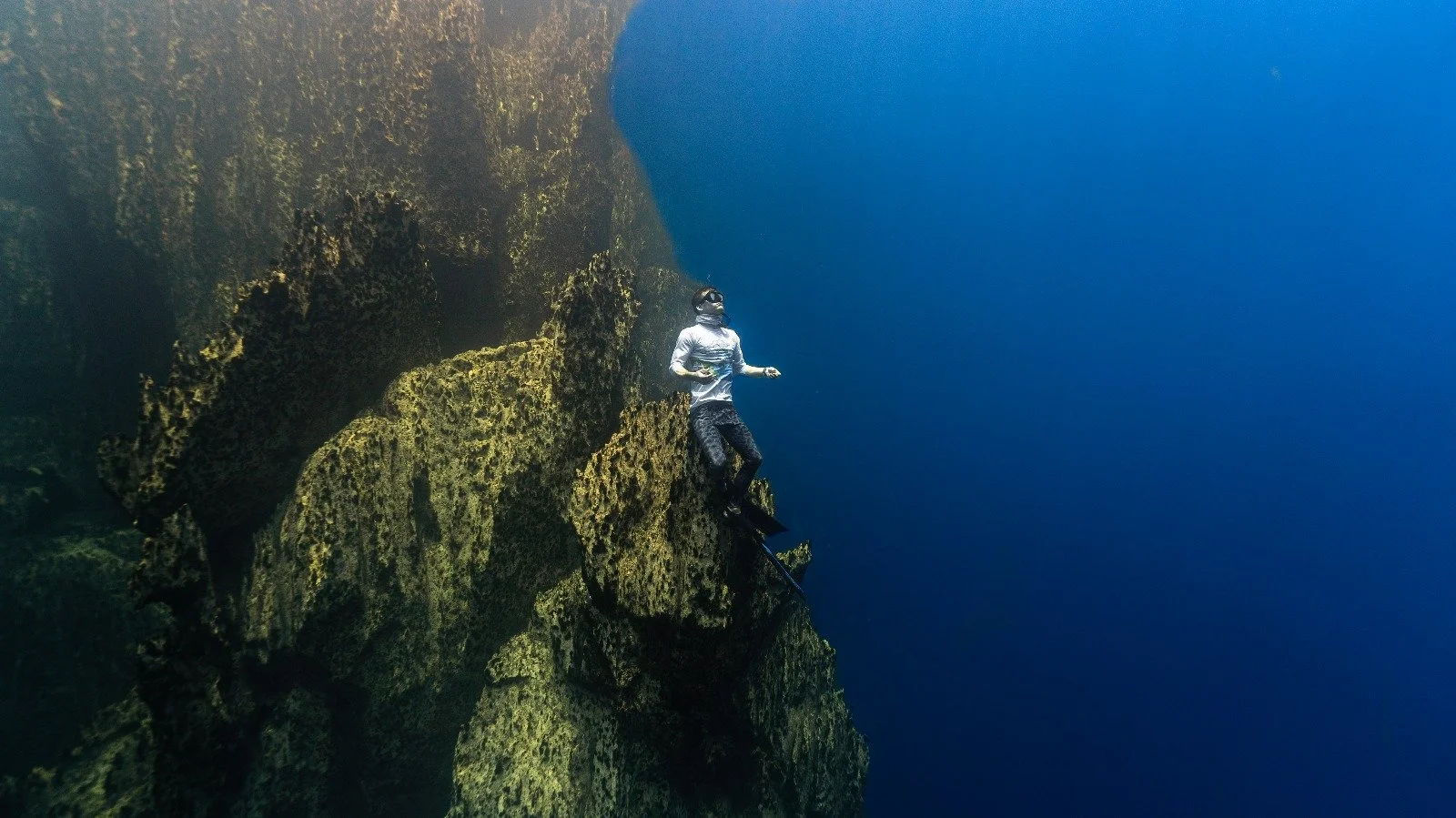 A freediver on a rocky underwater cliff, swimming in clear blue water.