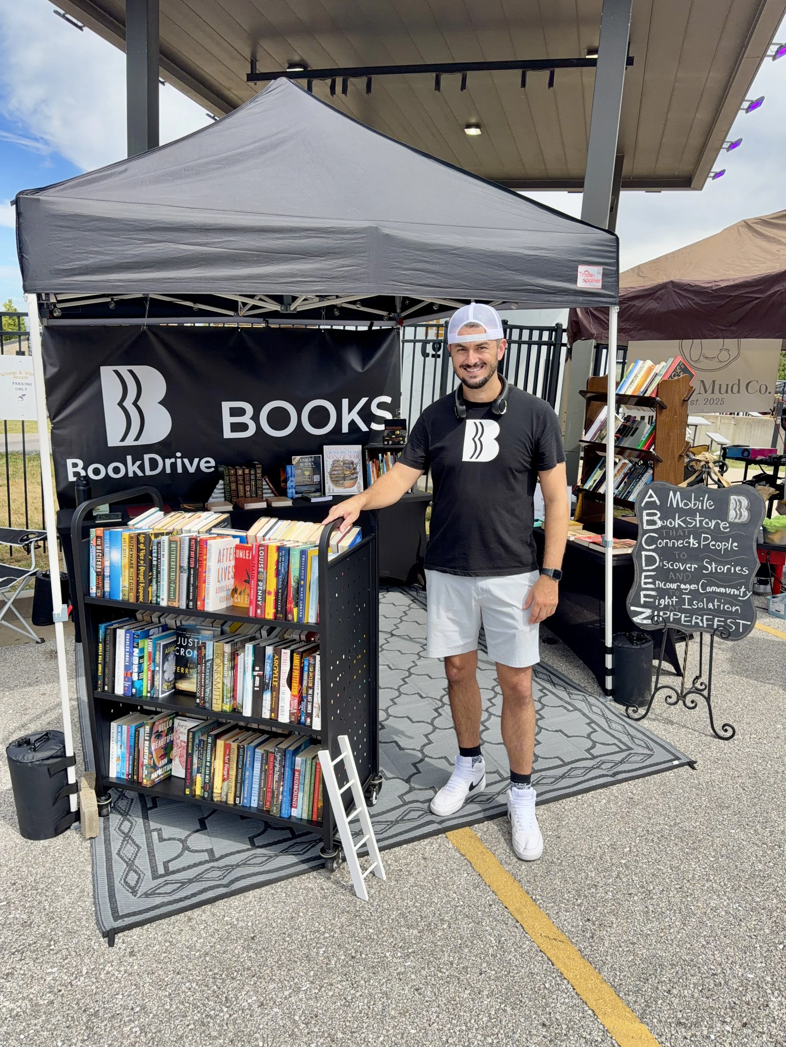 A man wearing a black t-shirt, white shorts, white sneakers, and a white baseball cap is smiling and standing at a bookstore booth set up outside. The booth has a black banner with white text that says 'BookDrive' and displays a large 'B' logo. The man is pointing at a cart filled with books.