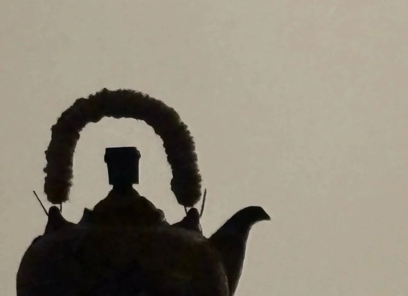 Silhouette of a teapot with a rounded handle, spout, and a small human-like figure sitting on the lid, creating a whimsical scene.