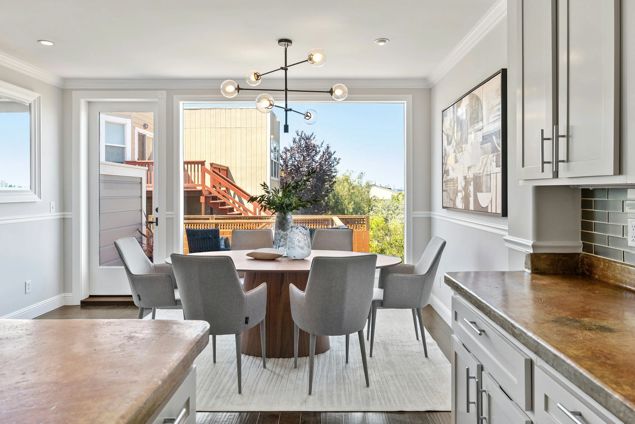 Modern dining area with a round table, gray chairs, and large windows showing an outdoor deck with stairs and trees.