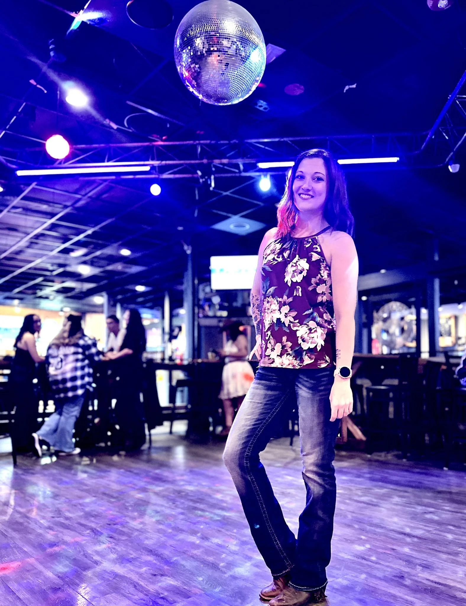 A smiling woman in a floral top and jeans standing on a dance floor in a nightclub with purple and blue lighting, a disco ball above, and other people in the background.