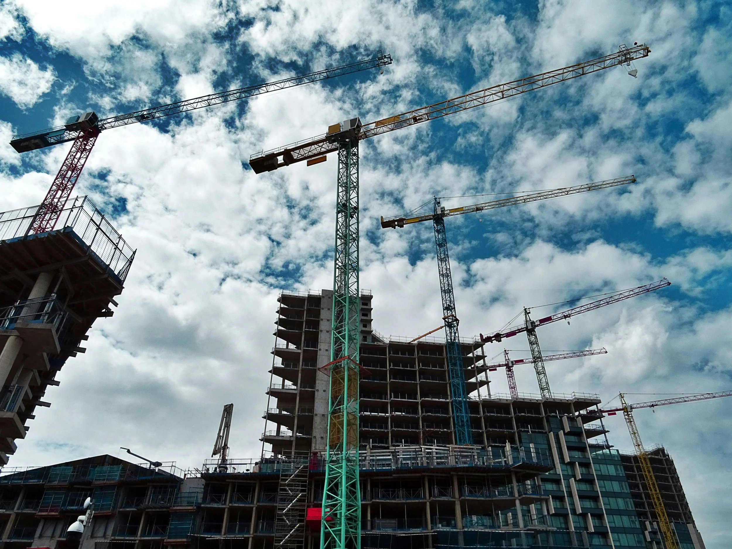 Construction site with multiple tower cranes and an unfinished multi-story building under a partly cloudy sky.