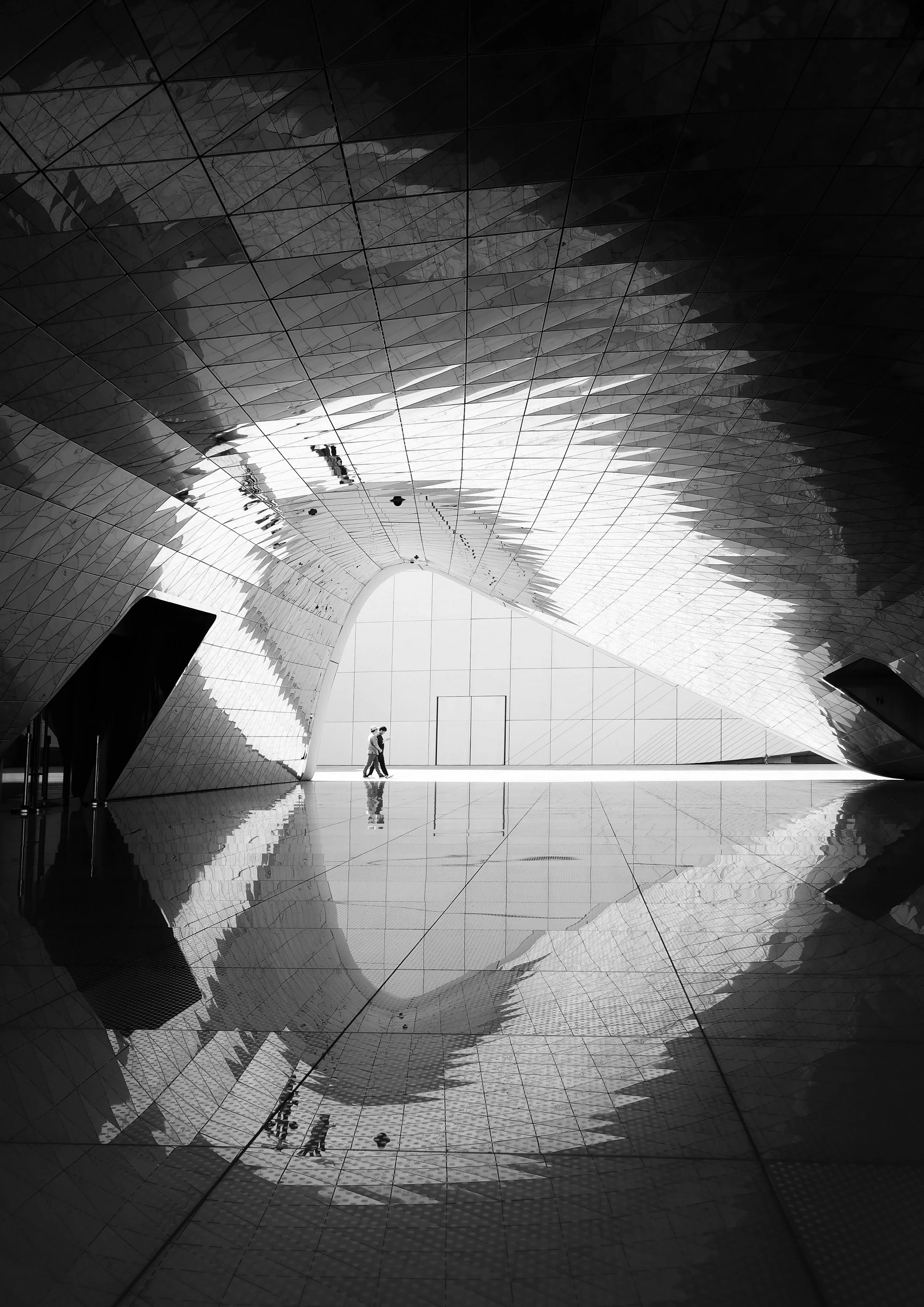 A black and white photo of a modern architectural structure with angular, reflective surfaces and a curved ceiling, featuring two people walking inside.