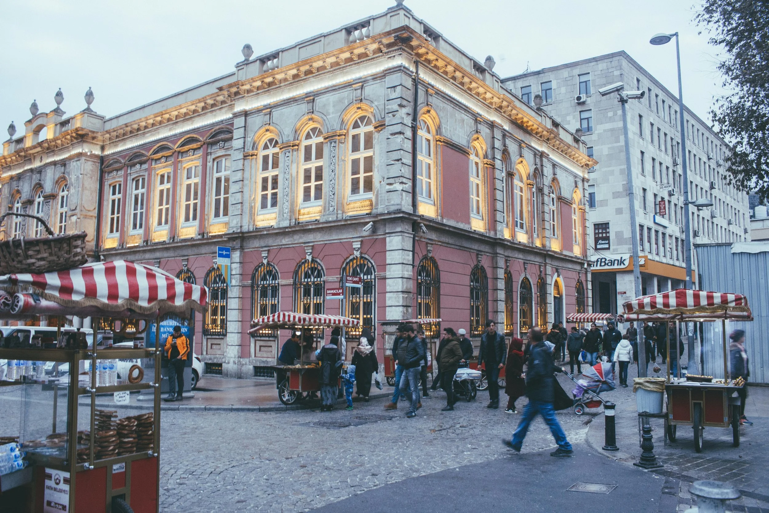 A busy city street corner with an ornate, historical building illuminated with decorative lights. Pedestrians walk past street vendors selling snacks and goods, with umbrellas providing shade, in an urban area with modern buildings nearby.