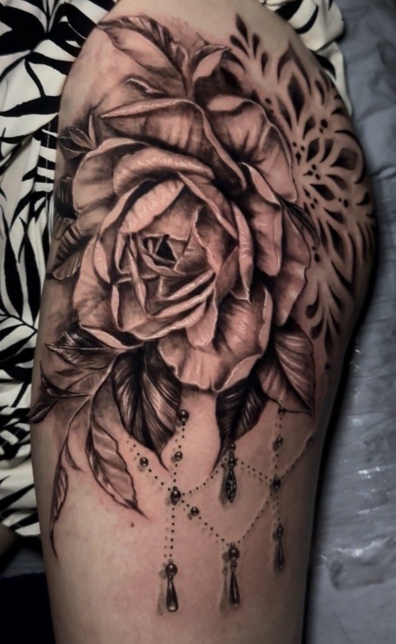 Black and gray tattoo of a detailed rose with leaves and a lace-like pattern, adorned with hanging beads and pendants, on a person's thigh.