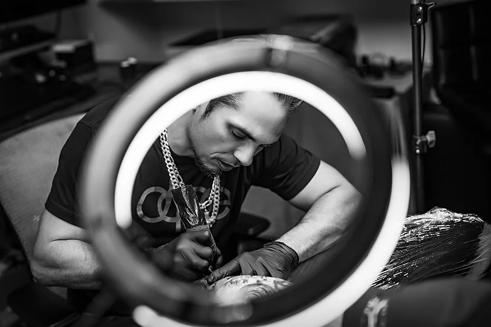 A man tattooing a person's arm in a tattoo studio, viewed through a ring light. Tattooist tattooer studio art body art construction design freehand free machine tattoo tattoos tattoo artist art professional private studio