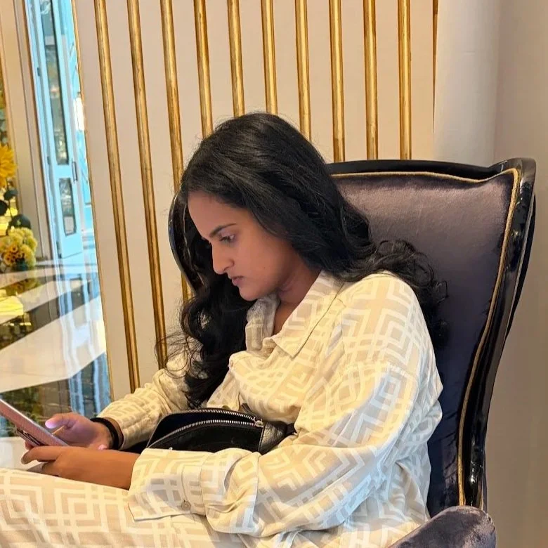 A young woman with long black hair, wearing a light patterned shirt, sitting in a black leather armchair and looking at her smartphone inside a decoratively paneled room.