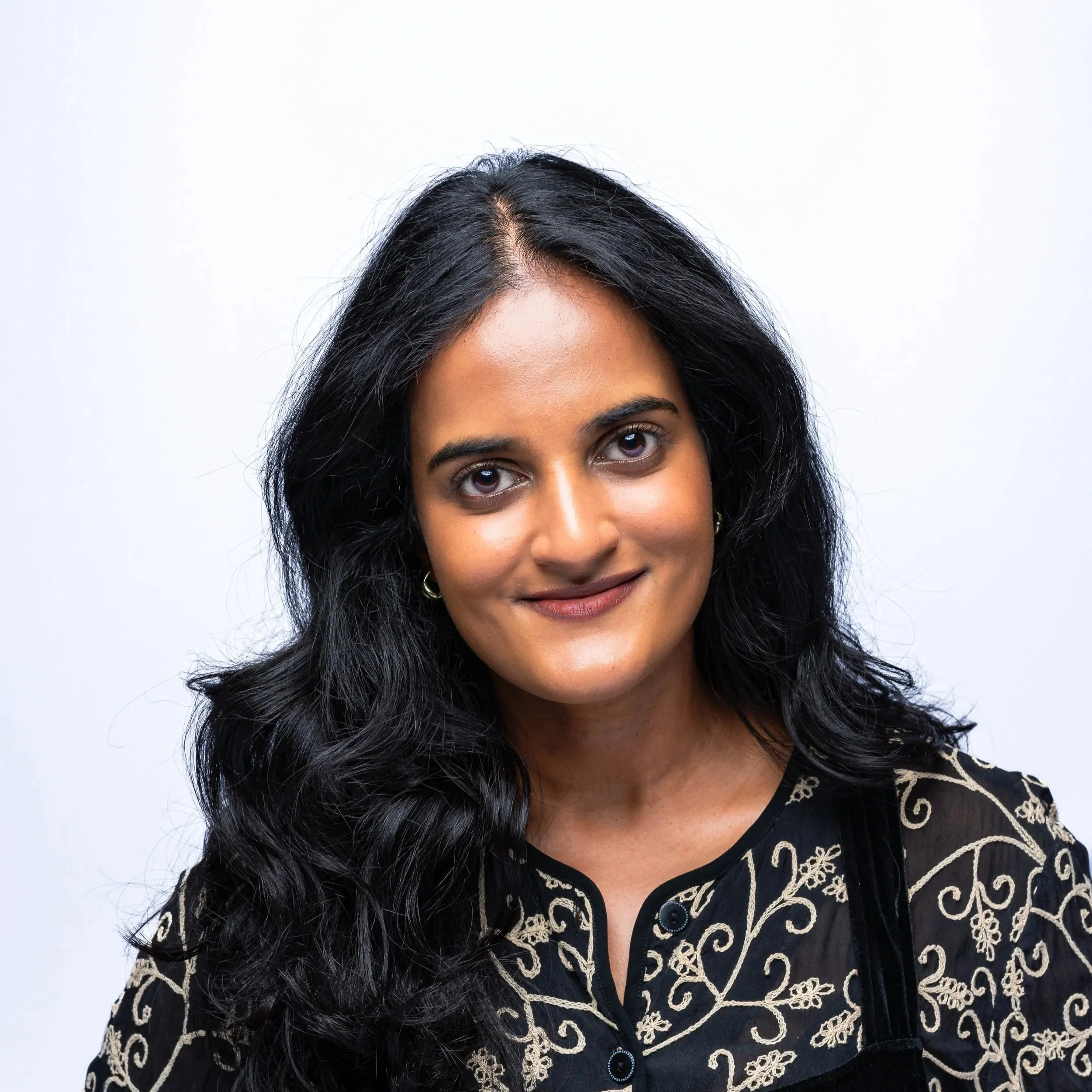 A woman with long, wavy black hair and brown eyes, smiling, wearing a black shirt with beige floral embroidery, standing against a plain white background.