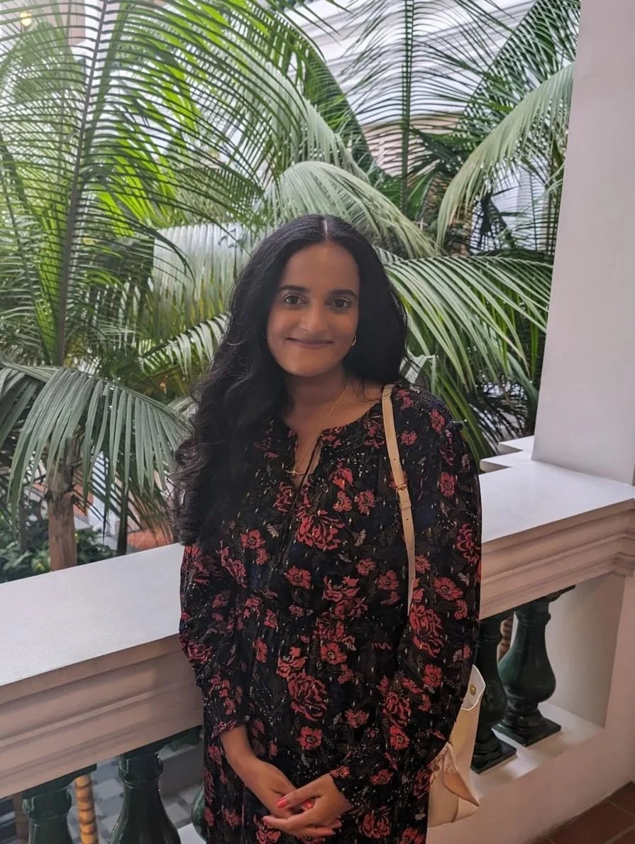 A woman with long black hair smiling at the camera, standing in front of lush green plants, wearing a long-sleeved floral dress and a beige shoulder bag.