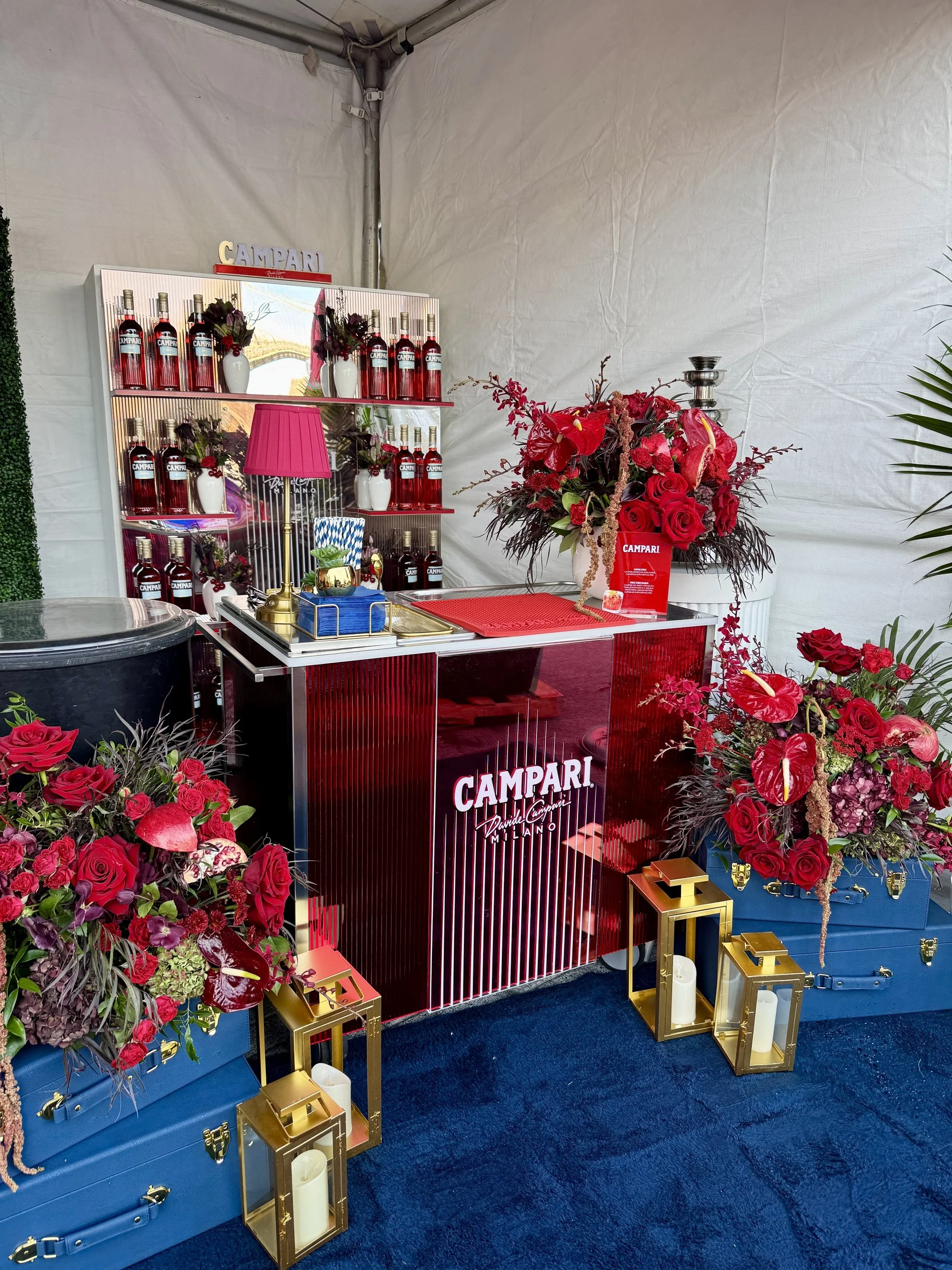 A promotional display for Campari featuring a red metallic counter with the Campari logo, surrounded by red floral arrangements, gold lanterns with candles, and various bottles of Campari on shelves.