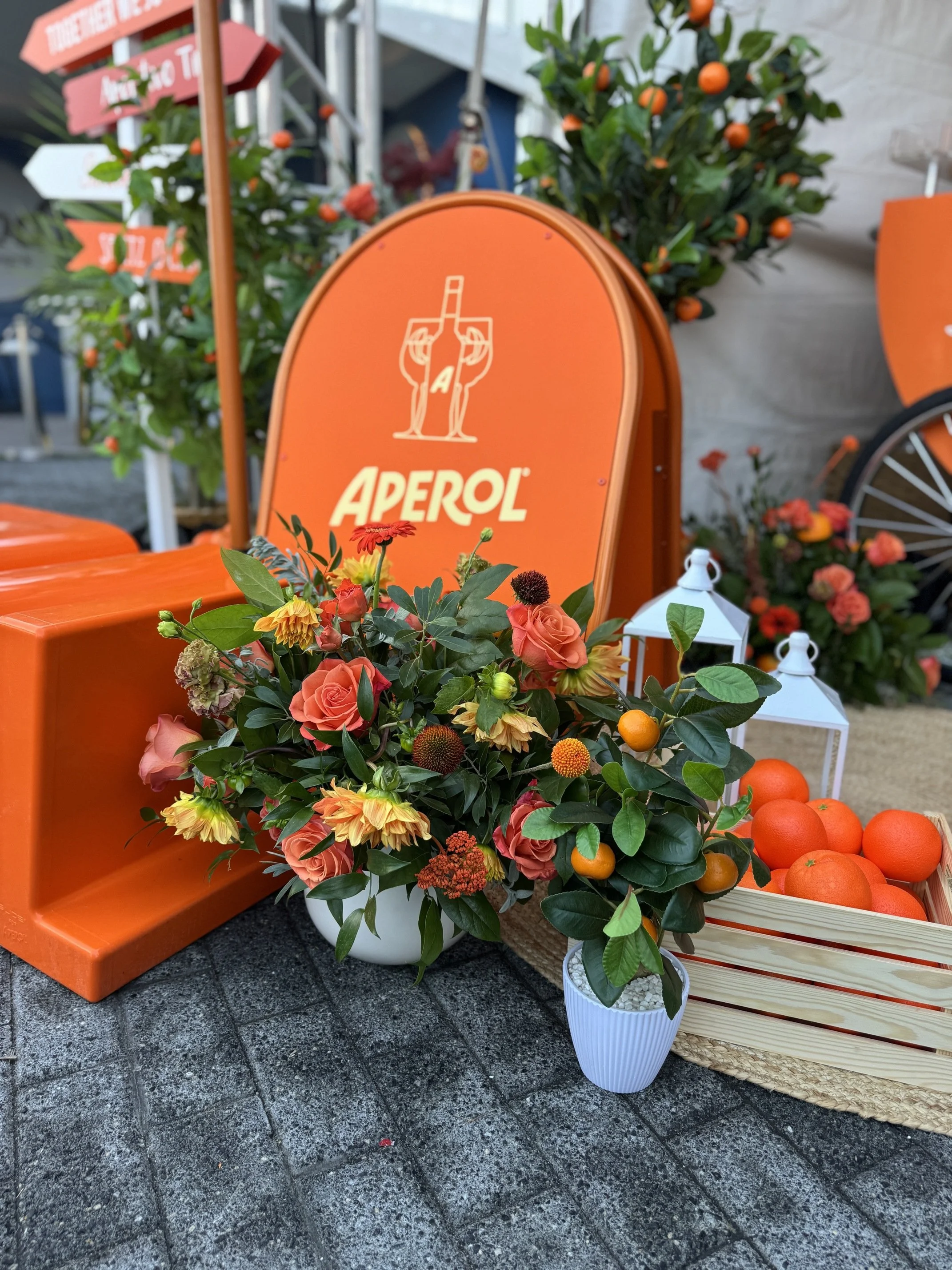 Display of flowers, oranges, and white lanterns arranged in front of an orange Aperol sign at an outdoor market or event.