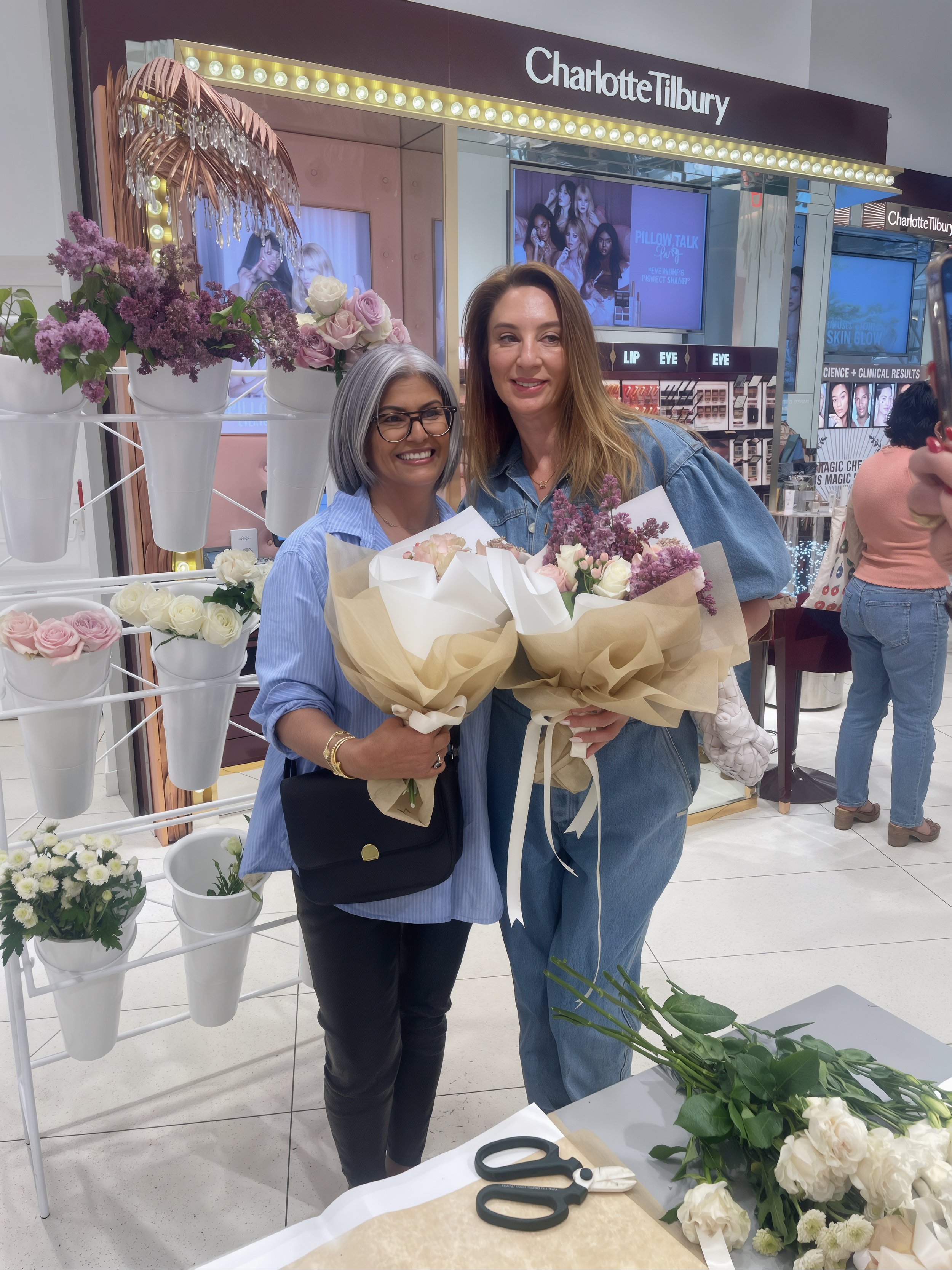 Two women holding bouquets of flowers smiling in a store with floral arrangements and a Charlotte Tilbury makeup display in the background.