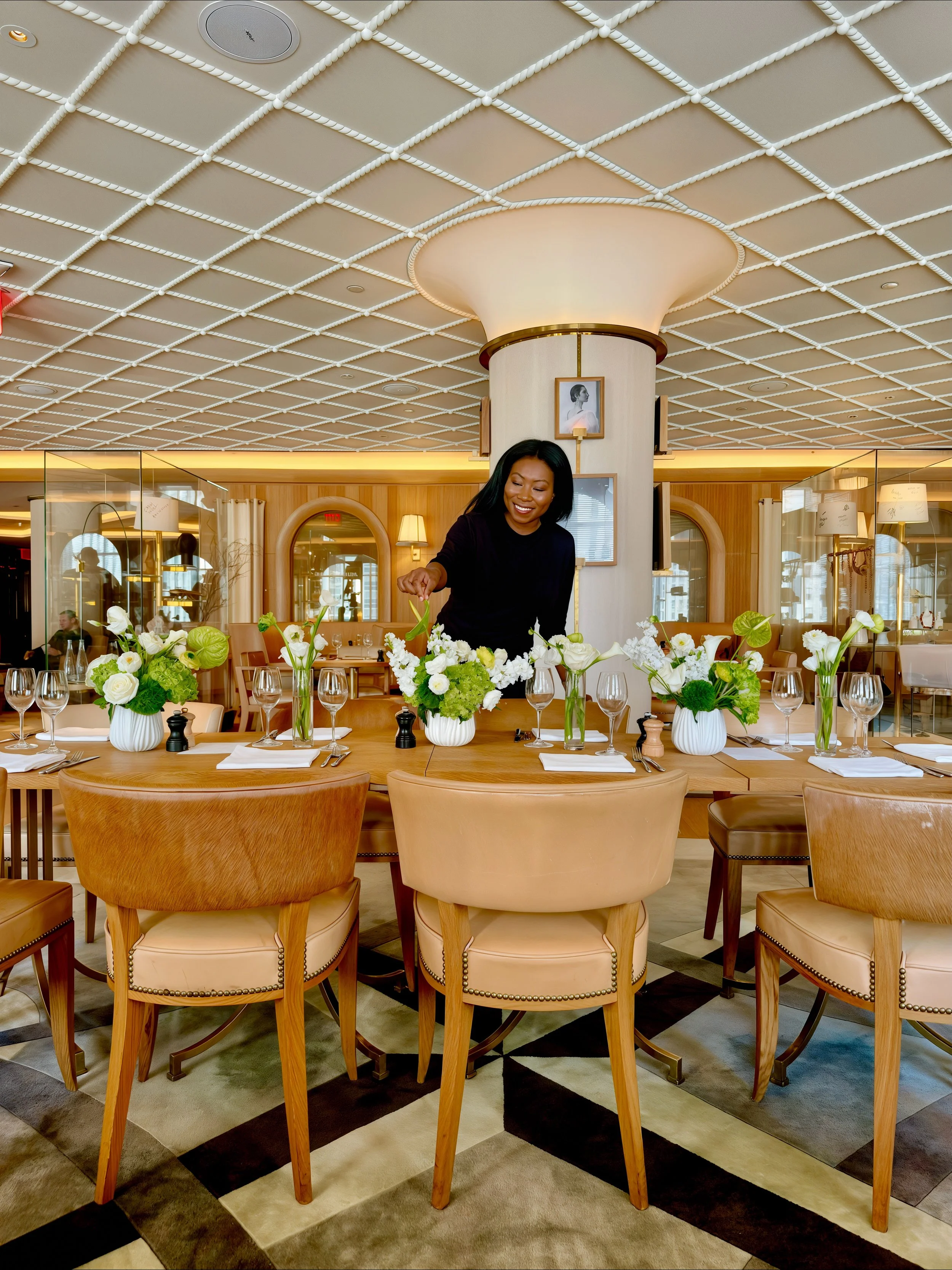 A woman arranging white flower centerpieces on a dining table in a well-lit, upscale restaurant or hotel dining room with wooden accents and decorative photographs on the wall.