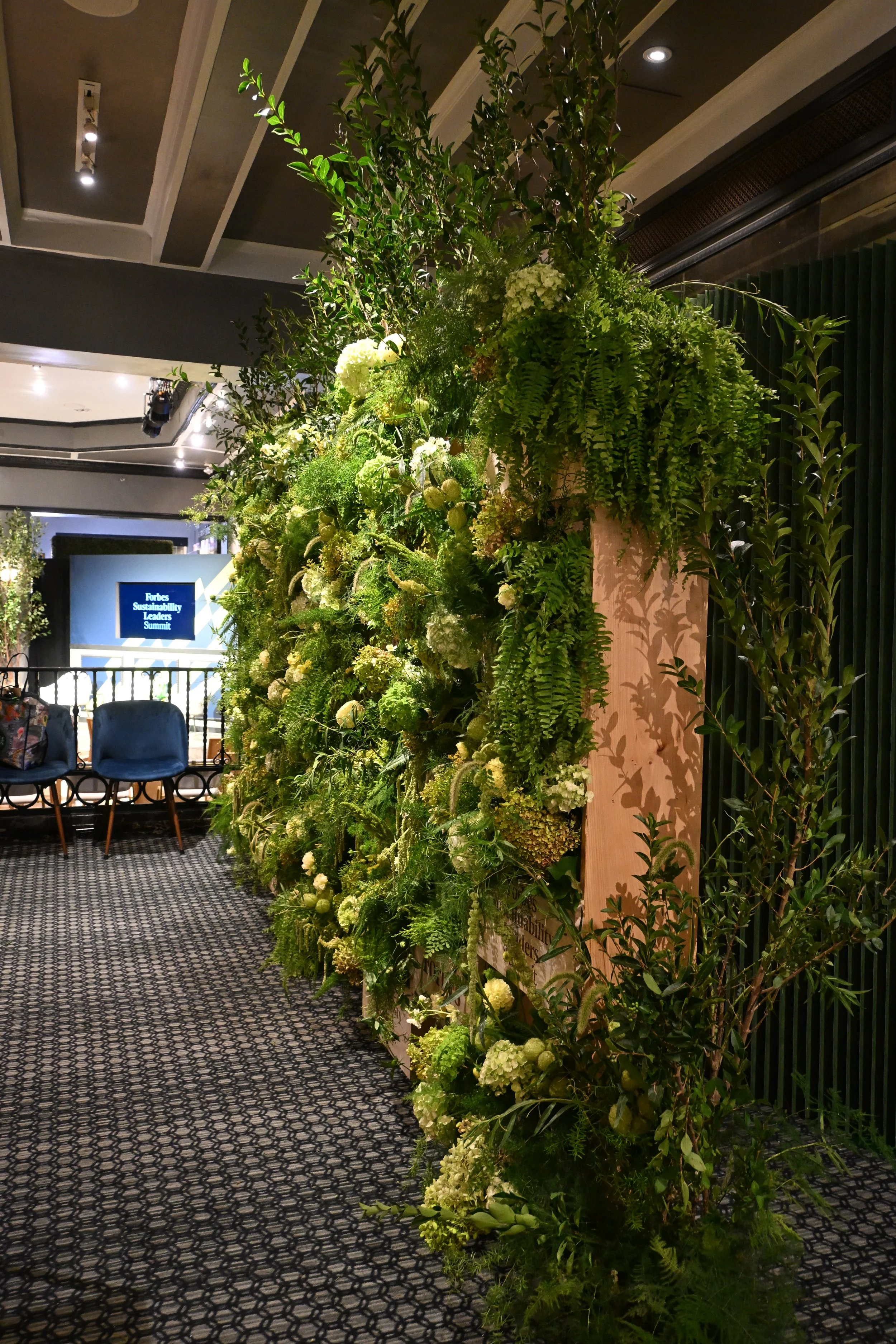 A large, vertical arrangement of various green plants and white flowers set against a wooden panel, located indoors at a conference or event, with chairs and a screen in the background.