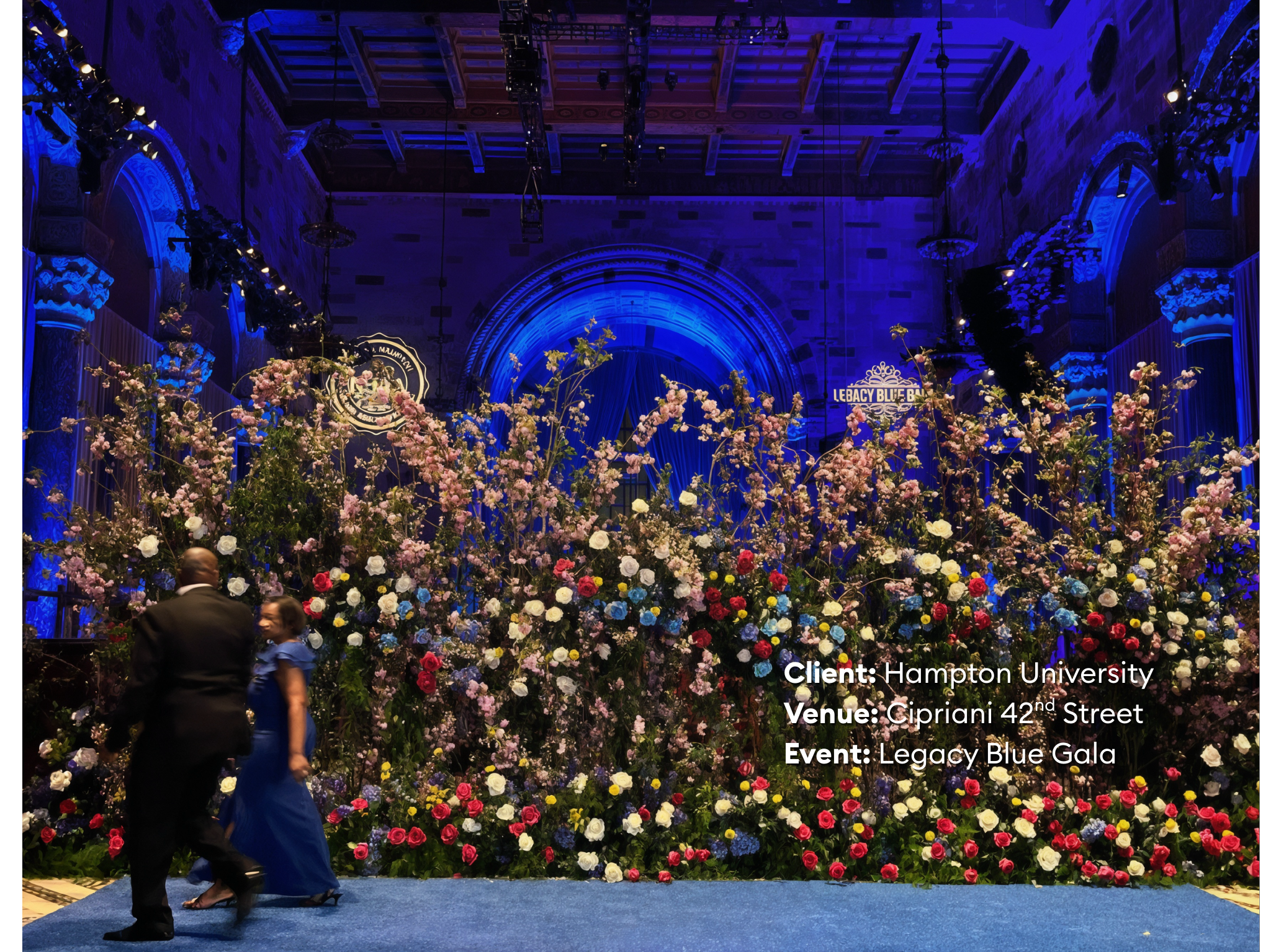 Elegant event in a grand hall decorated with a large arrangement of pink, white, red, and blue flowers. Two people, a man in a black suit and a woman in a blue dress, are dancing on a blue carpeted floor. The background features an arched stage with blue lighting and signs indicating the Legacy Blue Gala, with the client being Hampton University, held at Cipriani 42nd Street.