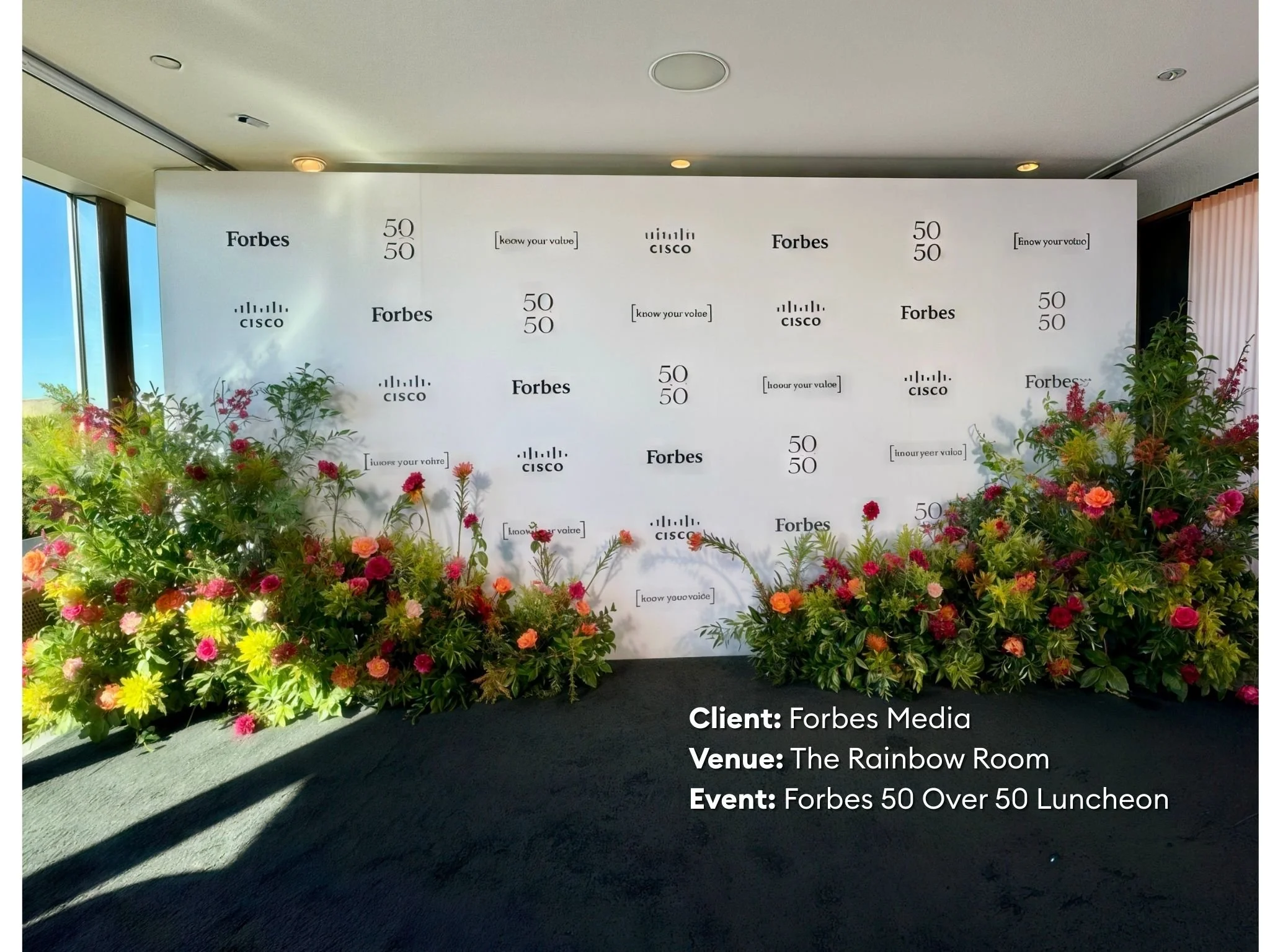 Event backdrop with fingerprints and Forbes logos, flanked by flower arrangements, at The Rainbow Room for a Forbes 50 Over 50 Luncheon.