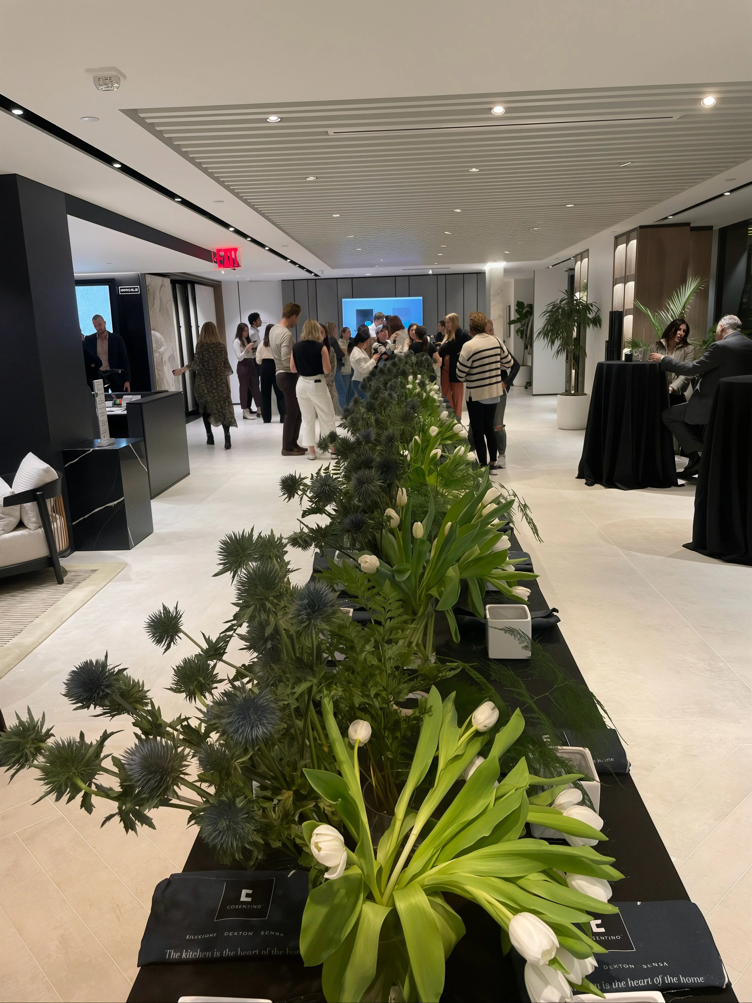 People gather around a display table with flowers in a modern, well-lit indoor space with plants and high ceiling.