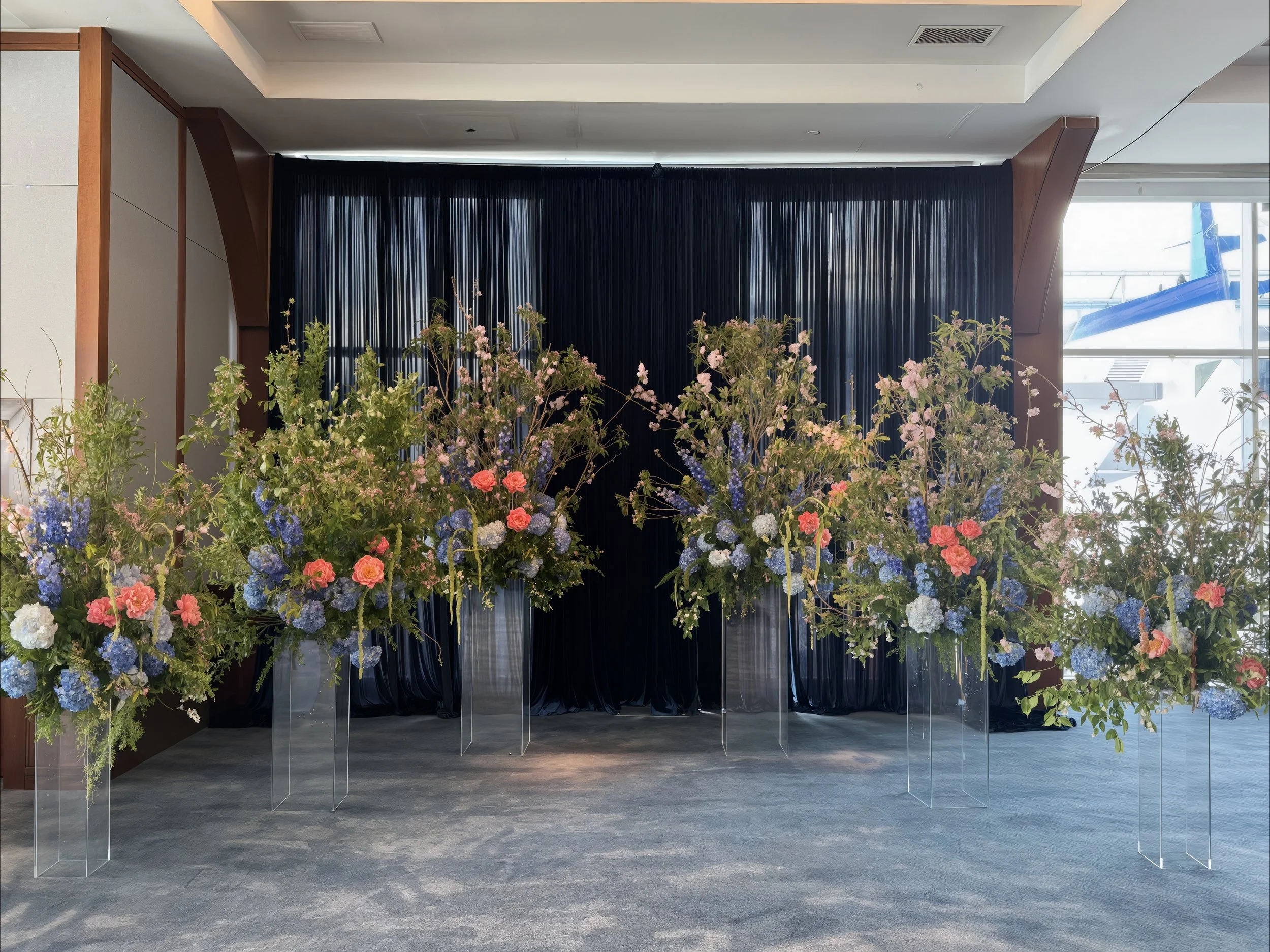 Elegant floral arrangements with pink roses, blue hydrangeas, and greenery on tall acrylic stands against a dark blue curtain backdrop, located in an airport terminal.