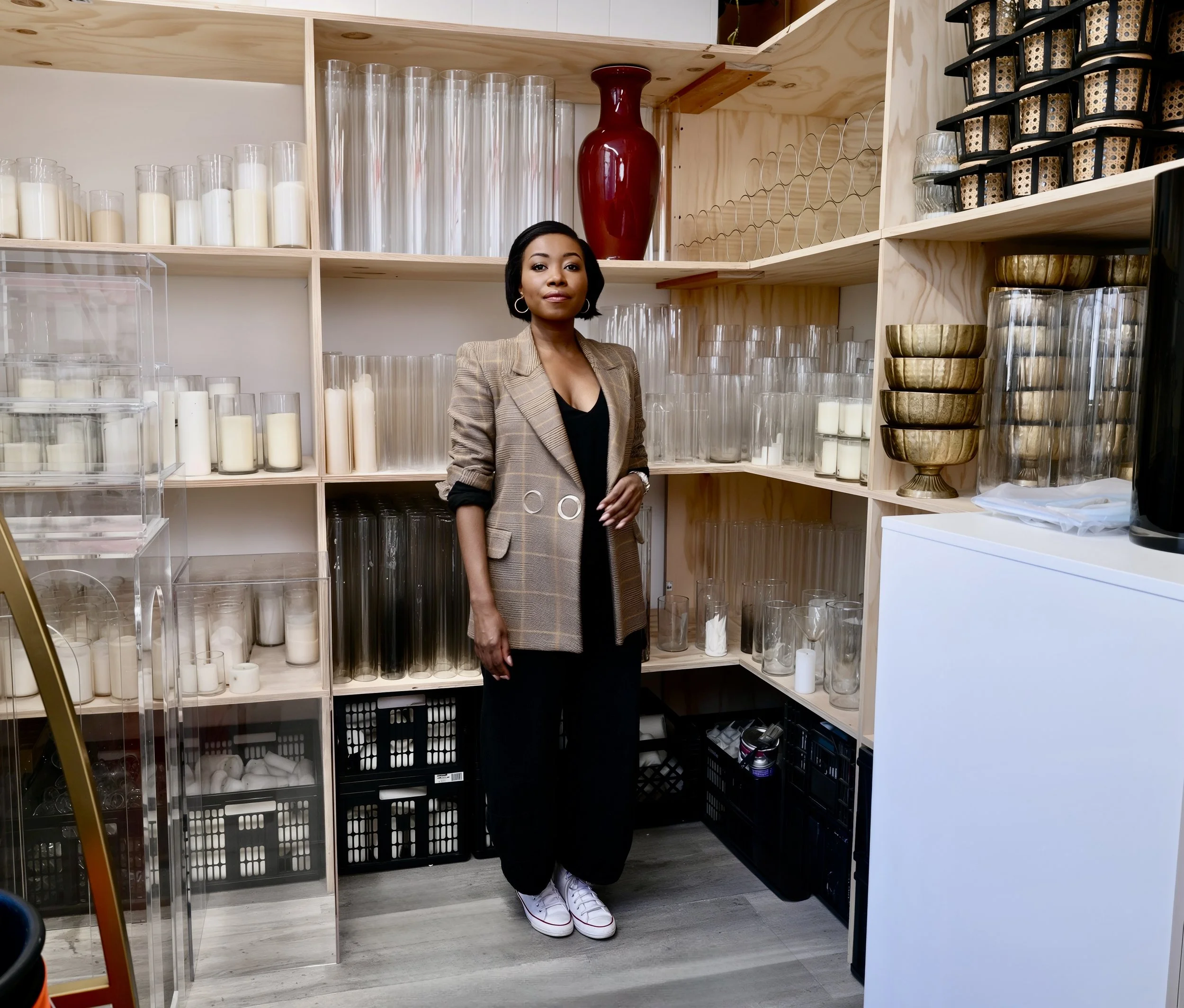 A woman standing in a room with wooden shelves filled with candles, glasses, and decorative bowls. She is wearing a plaid blazer over a black top, black pants, and white sneakers.