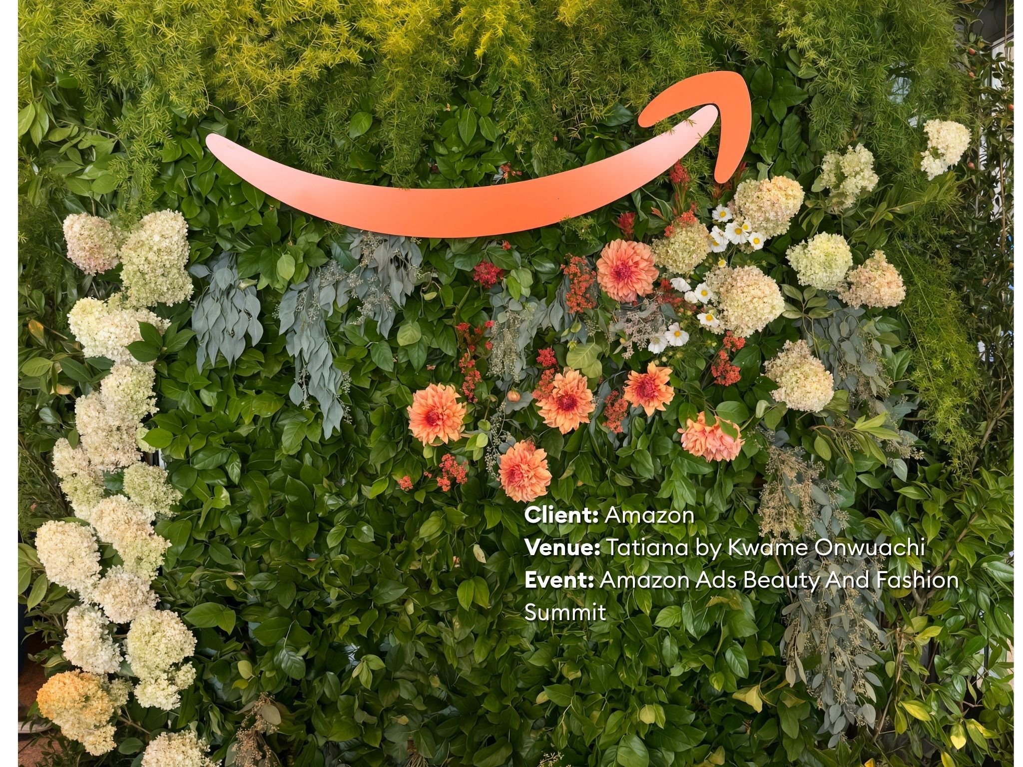 A lush green wall decorated with various flowers including white hydrangeas and peach dahlias, with an orange Amazon logo and event details overlayed.