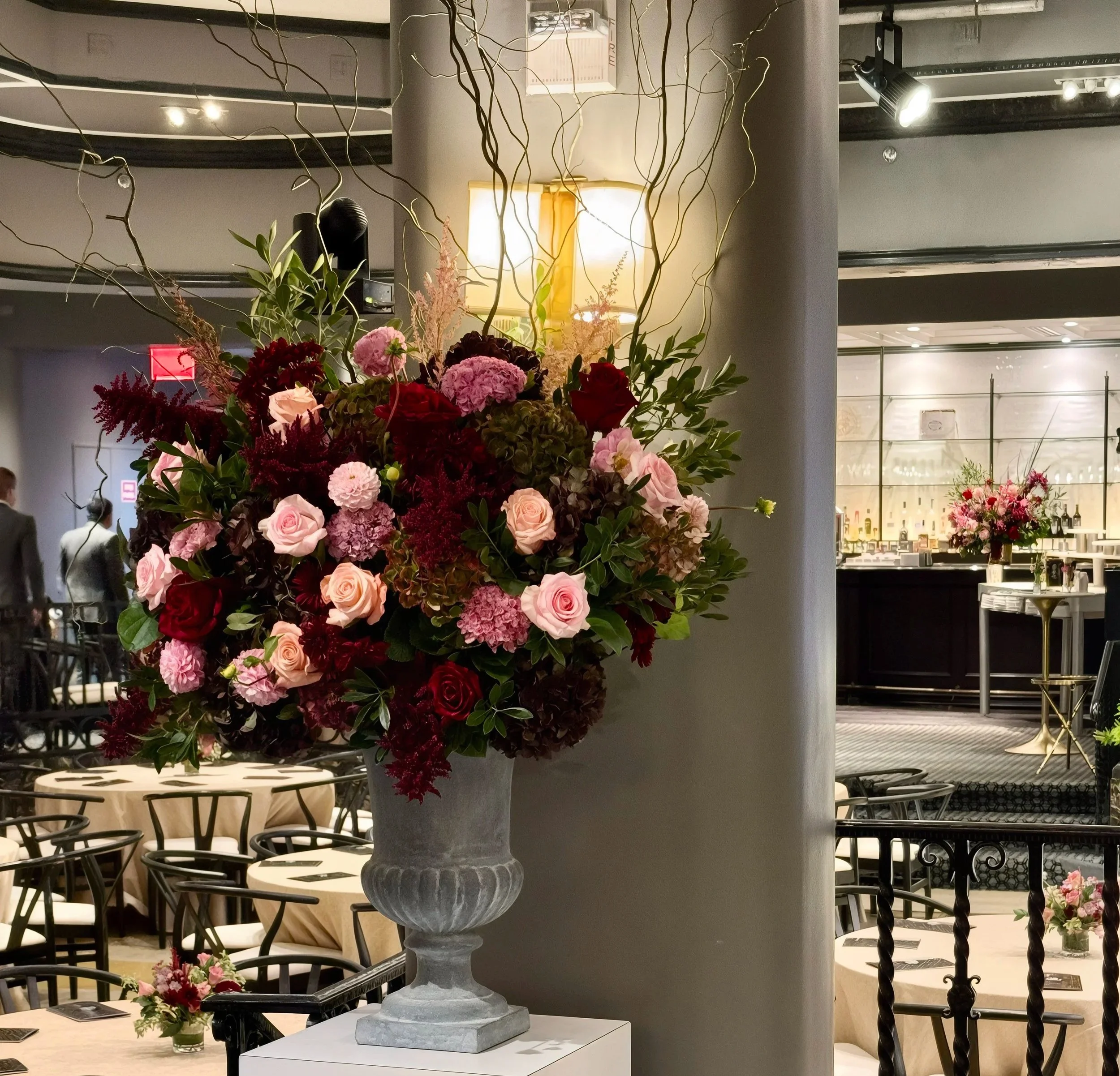 Large floral arrangement with pink, red, and purple flowers in a tall gray vase at a restaurant or event venue.