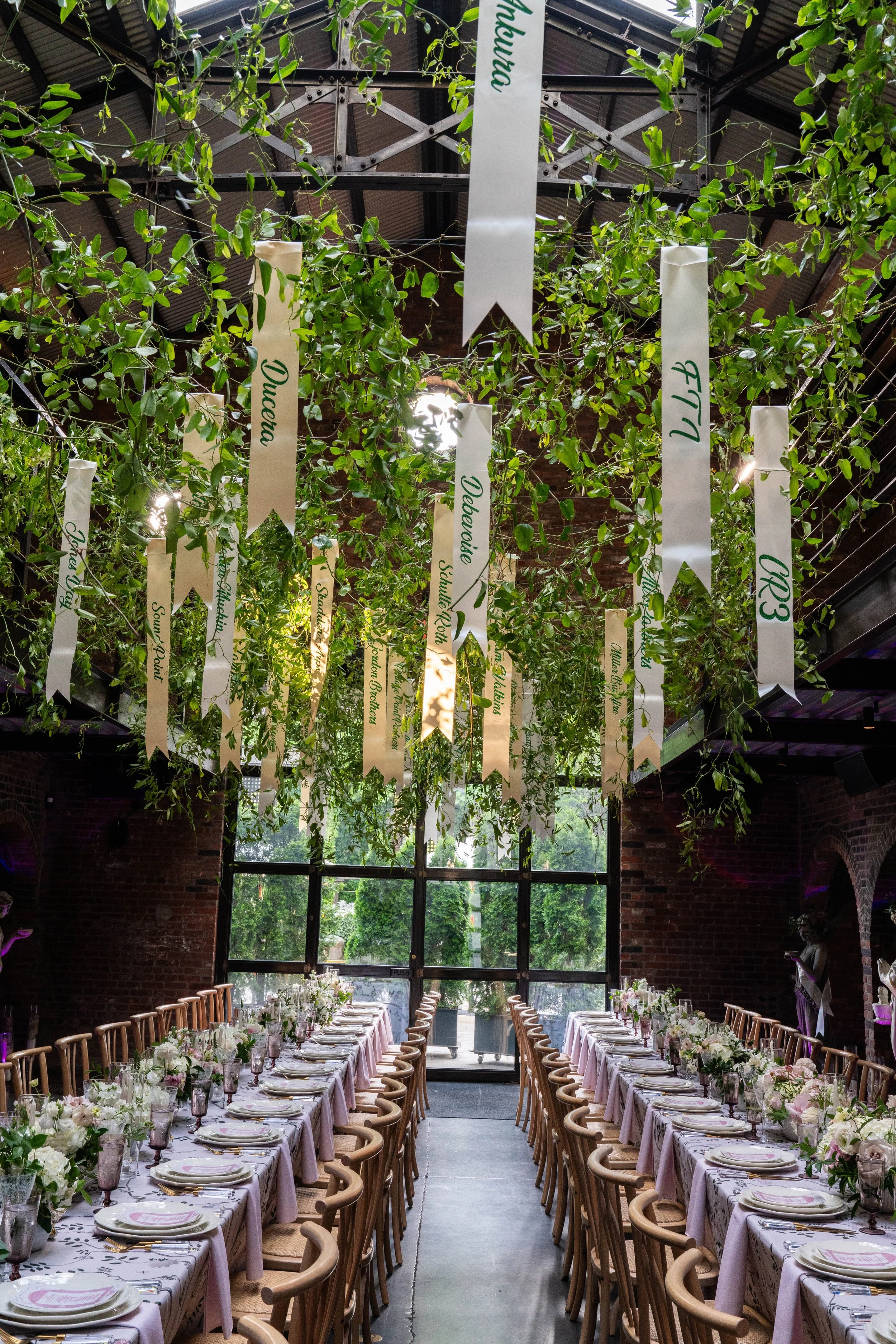 Long banquet tables set for a formal event or wedding reception, decorated with floral centerpieces, underneath hanging greenery and ribbons with labels, in a brick-walled industrial venue with large windows and natural light.