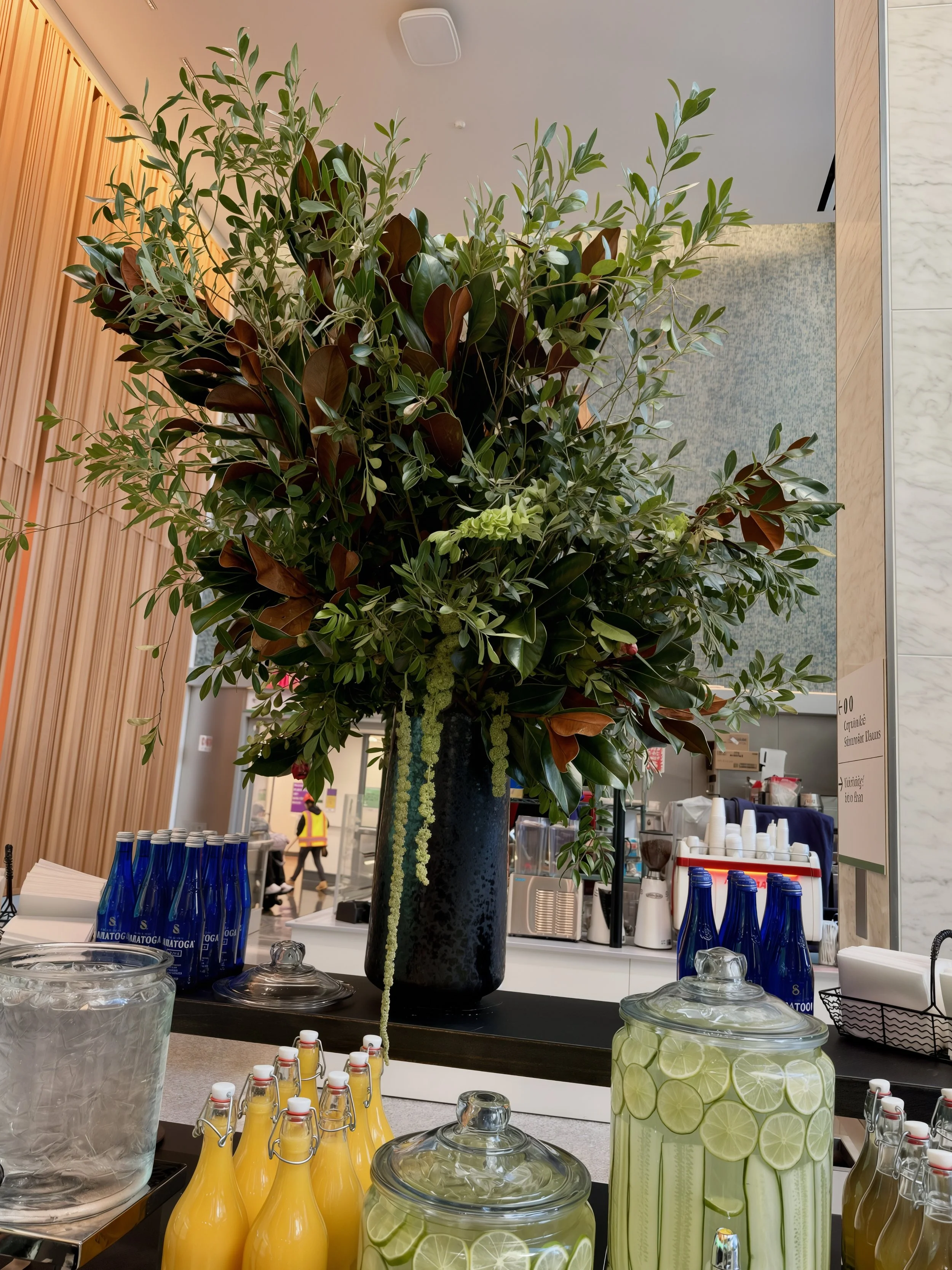 Large floral arrangement with green and brown foliage in a black vase, surrounded by bottles of yellow beverages and jars of lime-infused water, set on a table in a modern indoor setting.