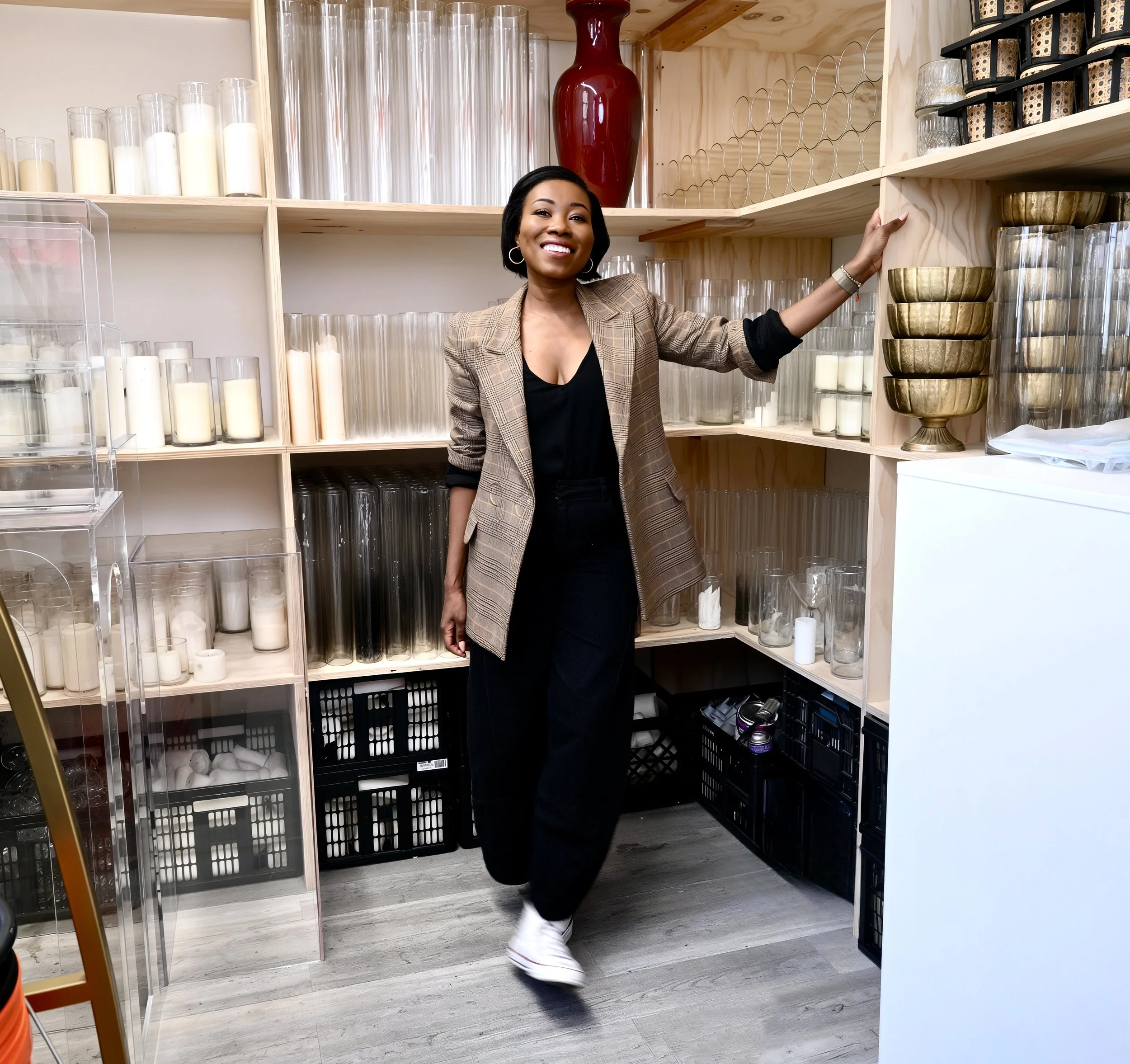A woman standing in front of wooden shelves filled with candles, vases, and decorative objects in a store or storage room.