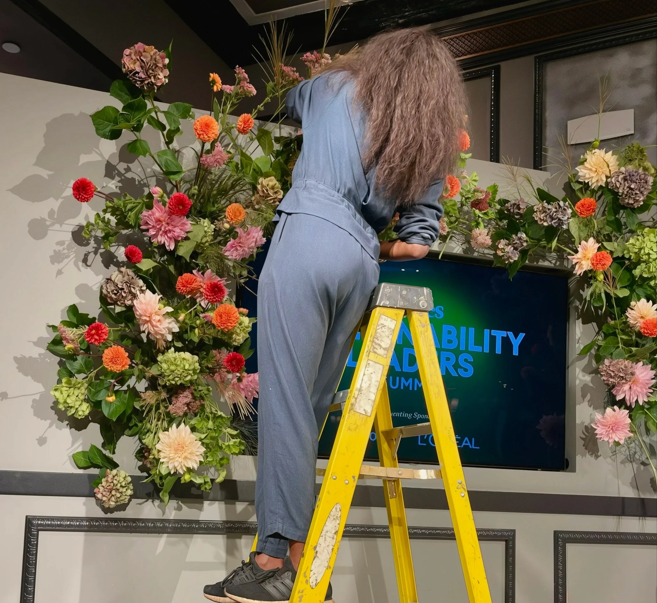Person standing on a yellow ladder arranging pink, orange, and purple flowers in a floral display behind a digital screen.