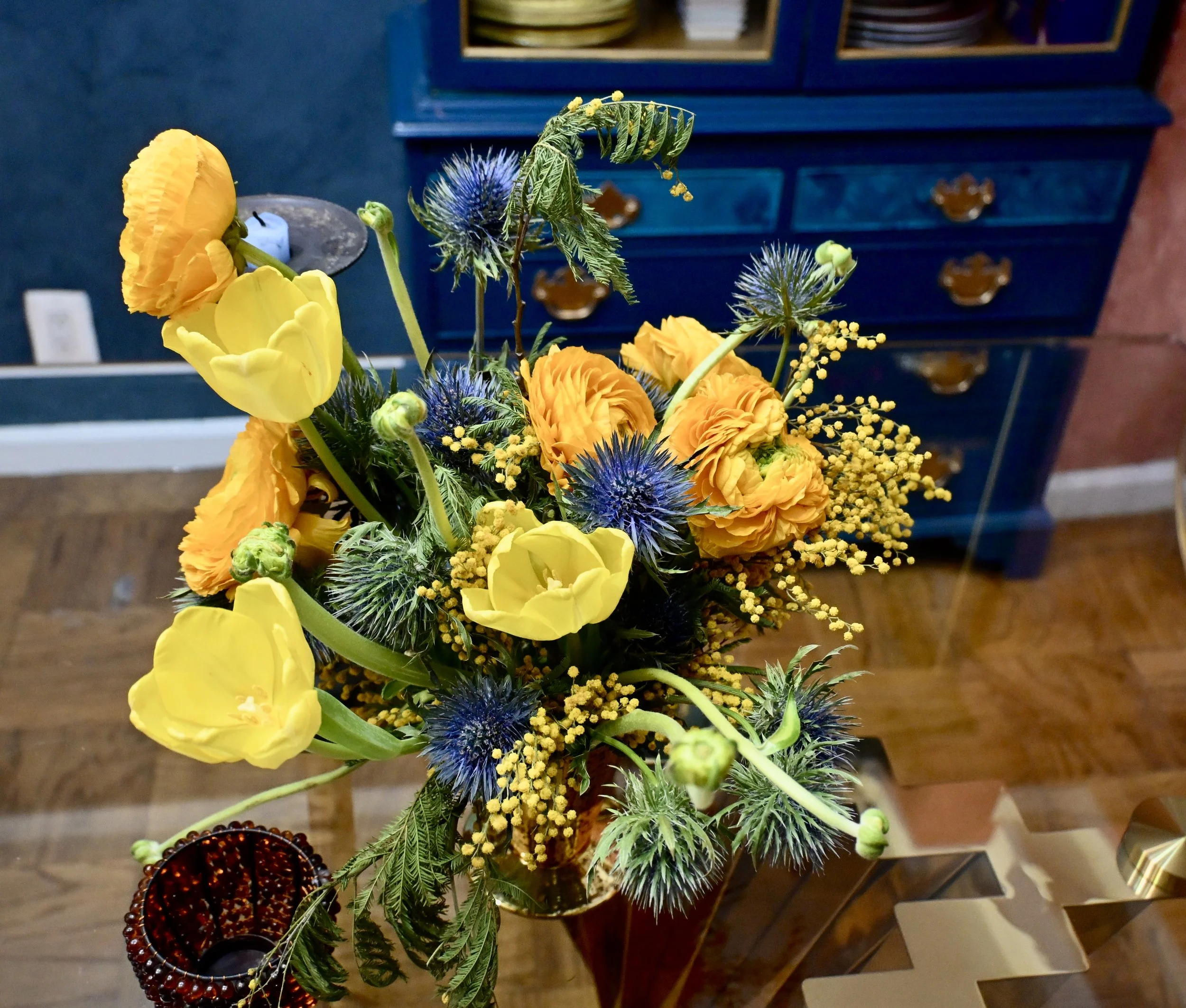 A colorful flower bouquet with yellow and orange flowers, blue thistle blooms, and yellow sprigs, placed on a wooden table next to a red glass candle holder, with a blue dresser in the background.