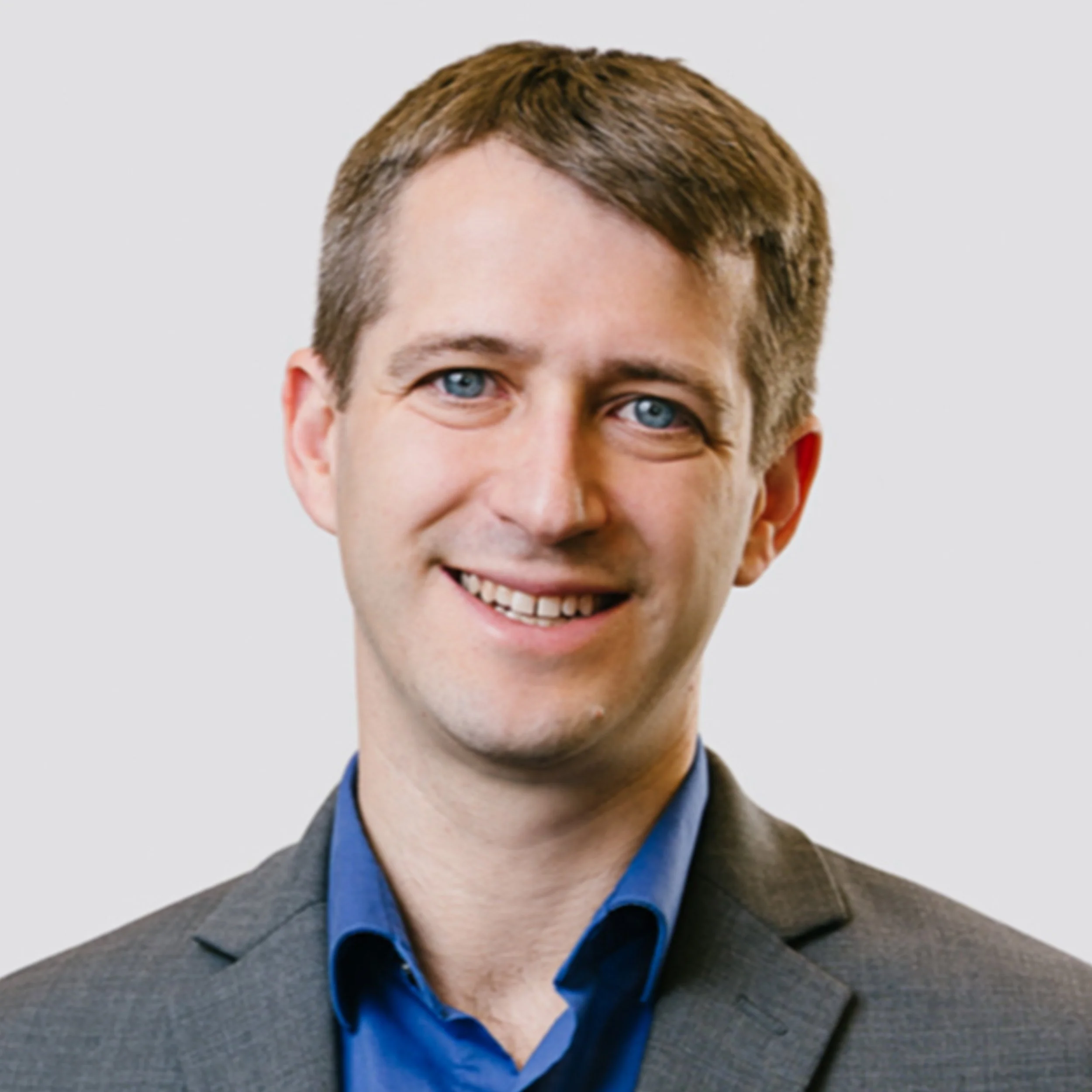 Portrait of a smiling man with short brown hair, blue eyes, wearing a gray suit and blue shirt against a plain light background.
